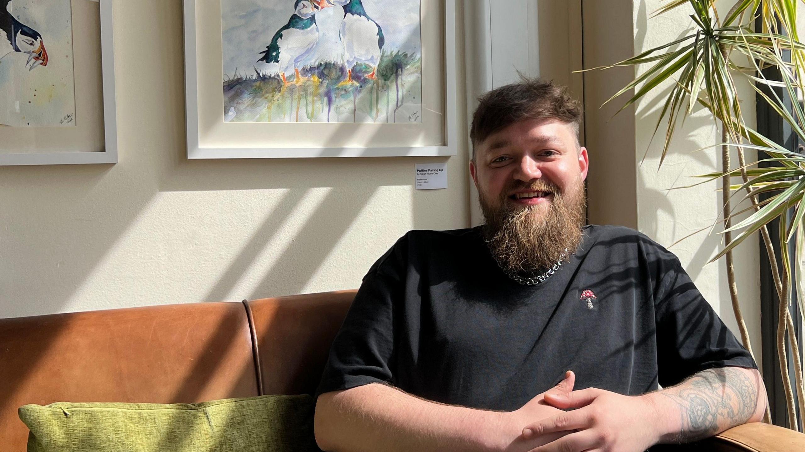 Mr Kabzinski is sitting on a brown couch, with green pillows, next to a window on a sunny day. He is wearing a black top and silver chain. His hands are clasped in front of him. Mr Kabzinski has brown hair, a long beard and is smiling at the camera.