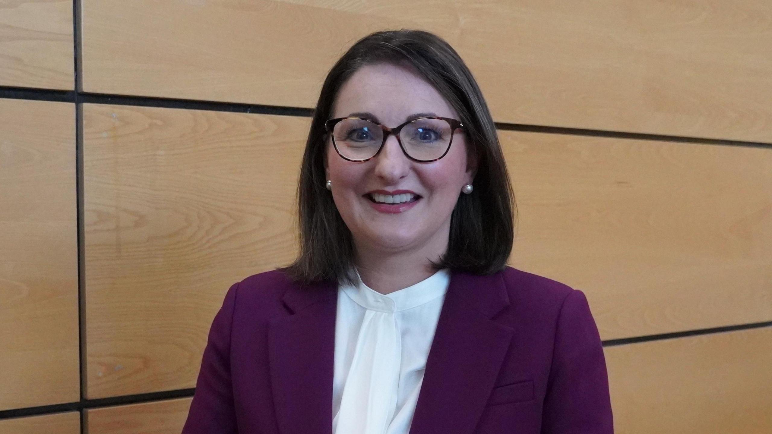 Ema Cubitt standing in front of a wooden panelled wall, smiling at the camera. She has shoulder-length dark hair and glasses and is wearing a purple suit jacket over a white blouse.