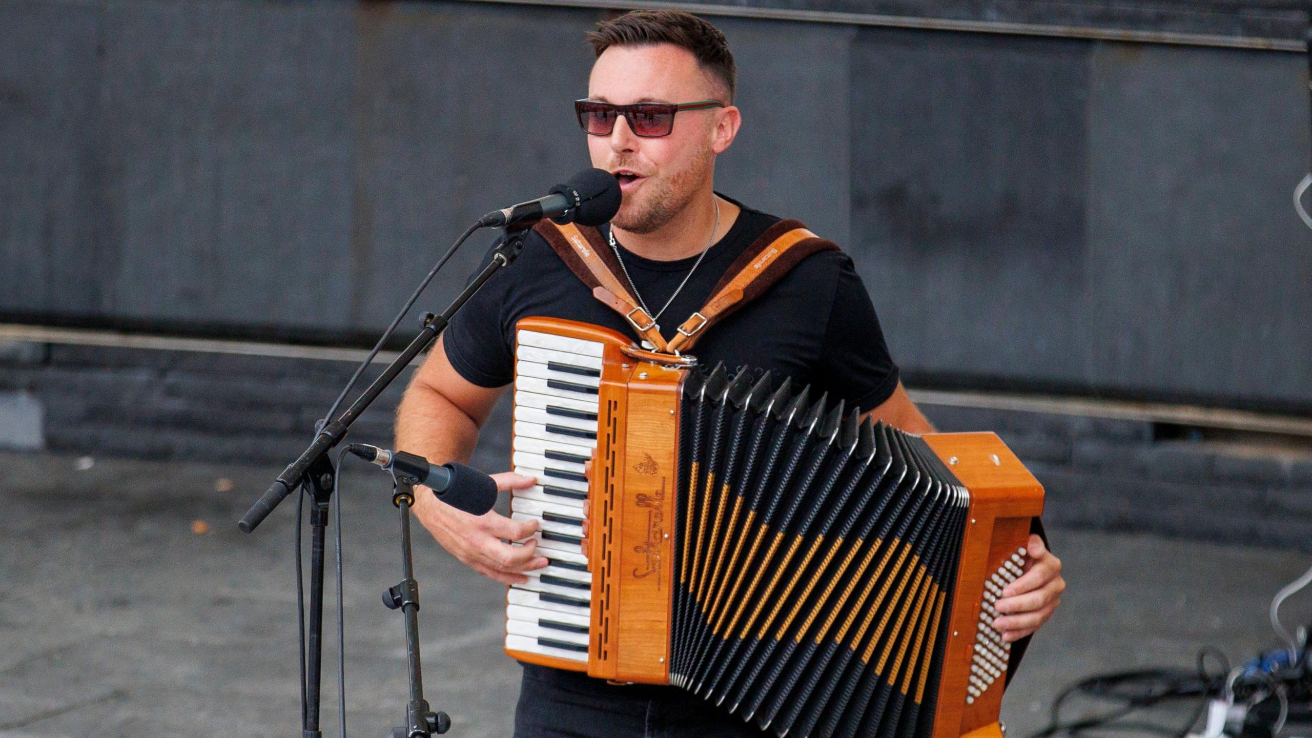 A young man with brown hair and brown glasses wearing a black t-shirt and silver necklace. He is playing the accordion in front of a microphone. 