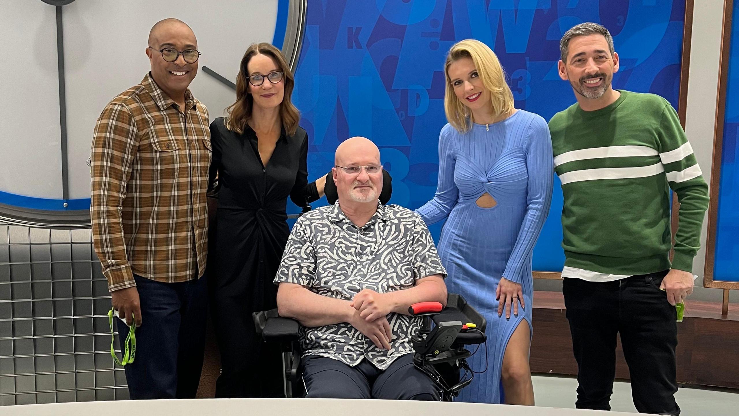 A man with a short, white beard is wearing a short-sleeved shirt and is sitting in a wheelchair. Either side of him are Countdown presenters and the other contestant. They are gathered on the set of the quiz show.