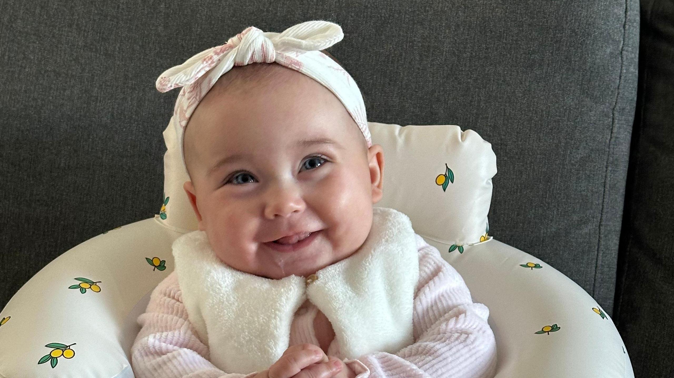 Baby Sophie McCracken smiling at the camera. She is a few months old and is wearing a pink long-sleeved top and a furry collar. She has a bow in her hair and is clasping her hands together.