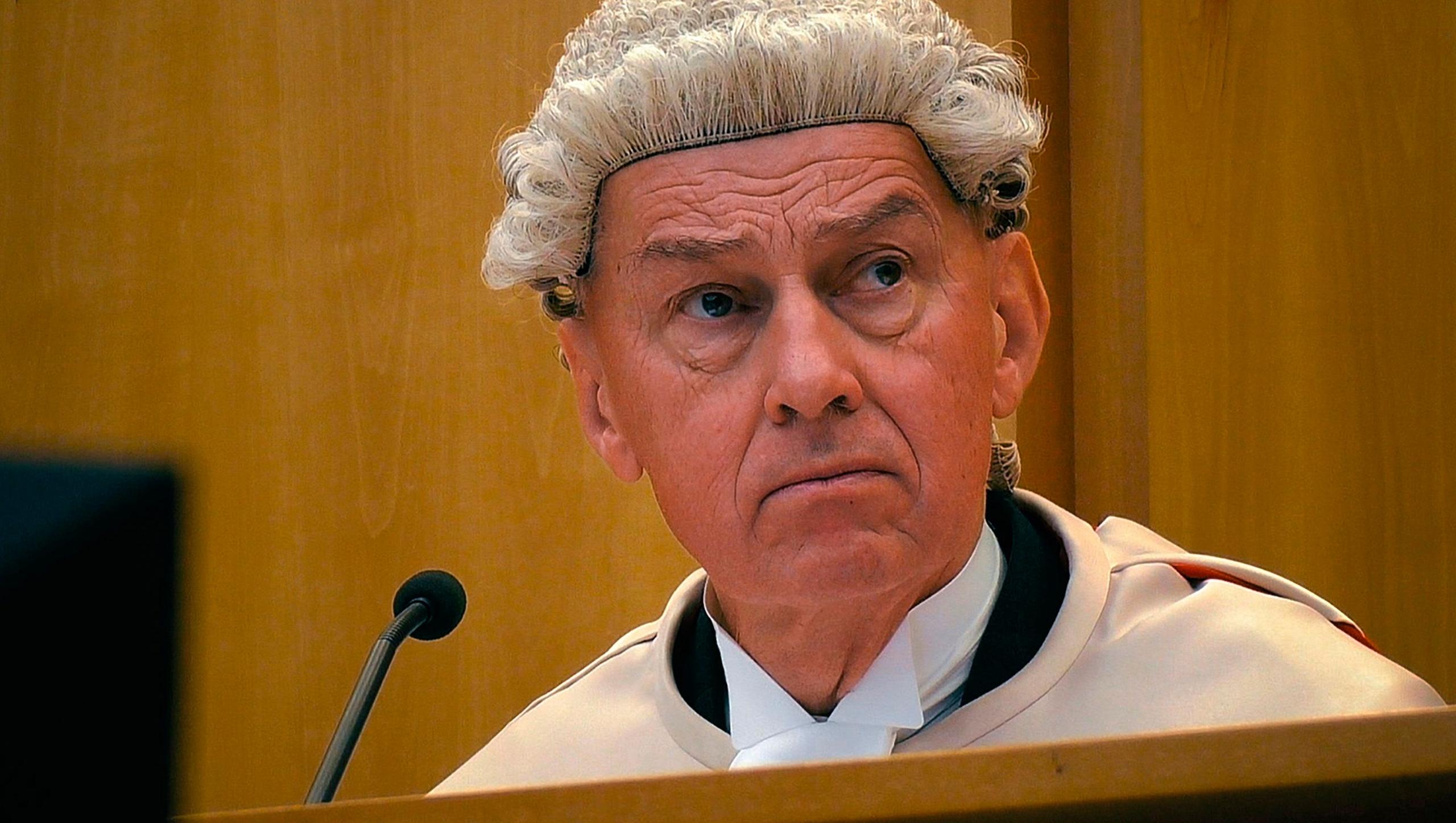 Photo of the judge in court. He is wearing his court robes and wig. He is sitting in front of a microphone and looking towards the court.