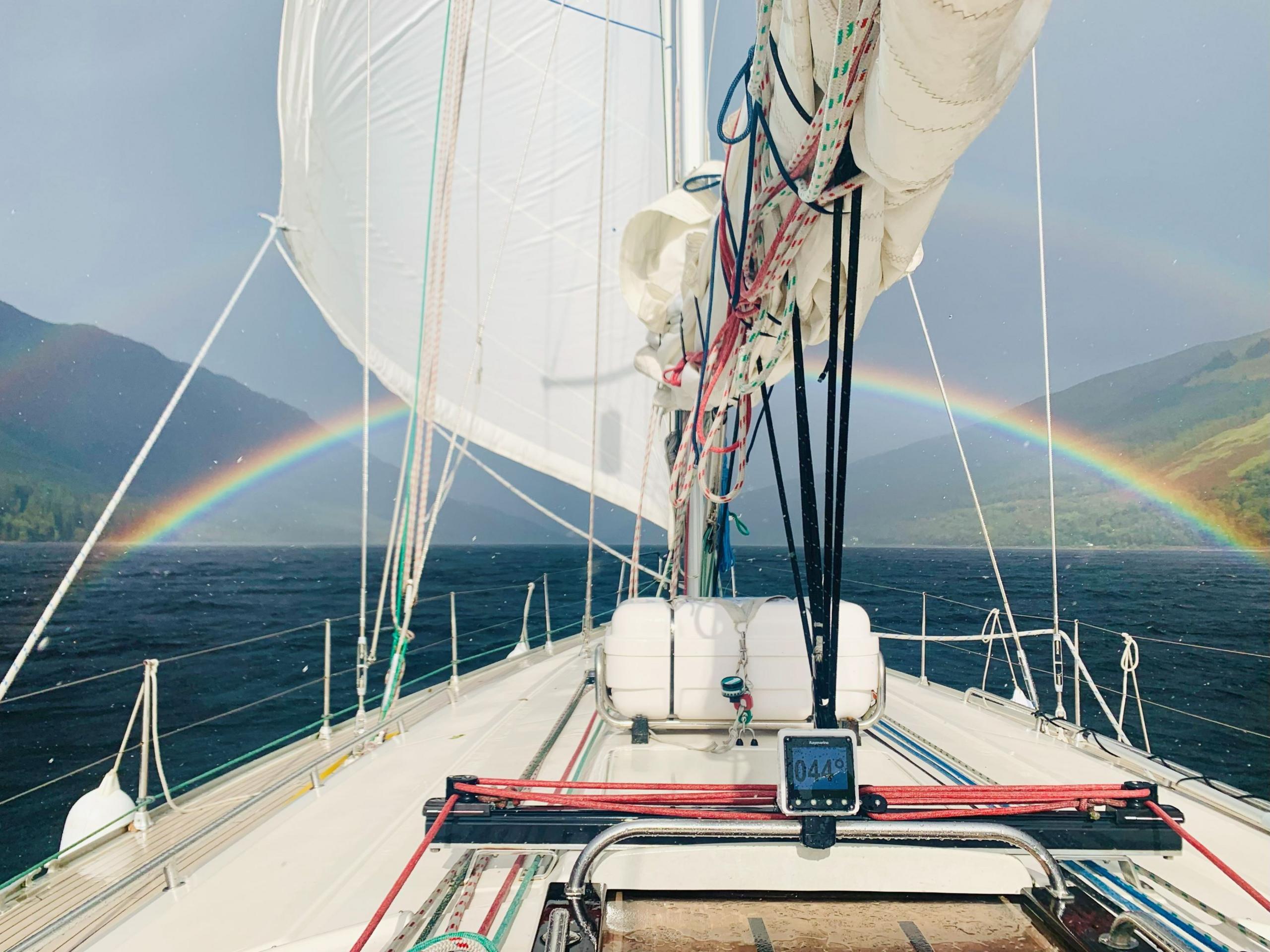 A view down to the bow of a sailing boat, masts, sails and ropes all visible, sailing through a loch with hills on wither side. In the sky on front is a beautiful double rainbow which arches over the bow of the boat.