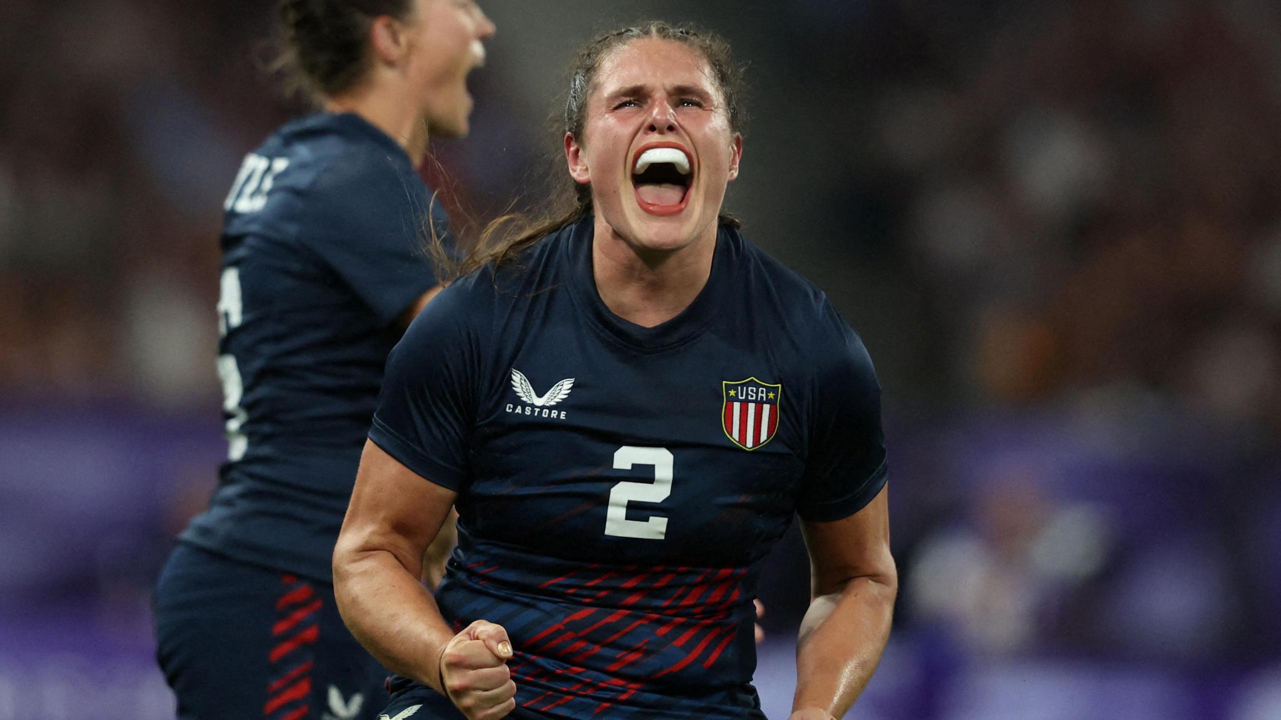 Ilona Maher playing for Team USA during the Paris Olympics. She wears the Team USA kit - a tight blue top with red stripes across the midriff and the US flag on her chest. She's pictured mid scream, her fists clenched. 