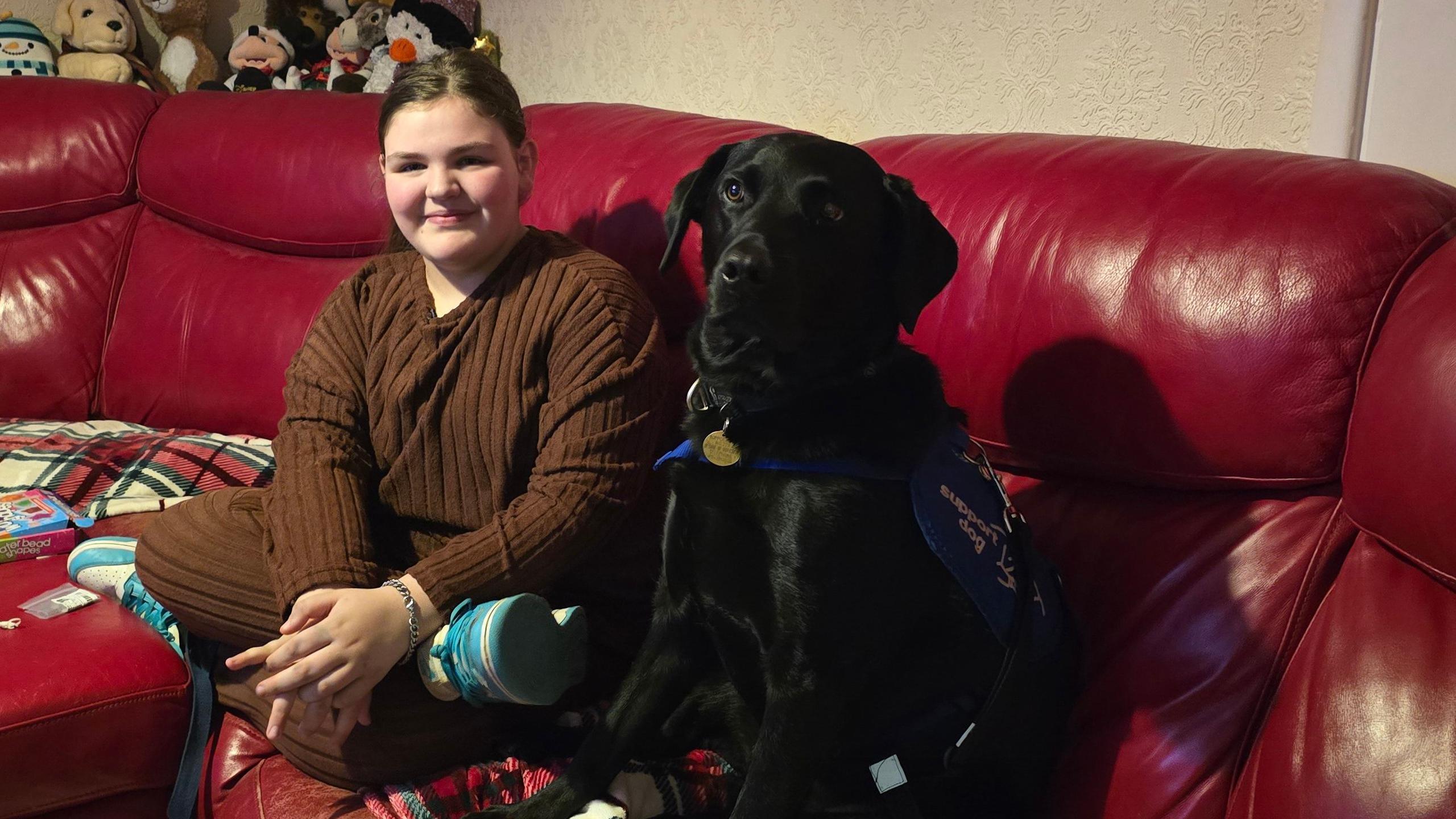 Scarlette, wearing a brown tracksuit, sits in her sitting room on a red sofa with her black Labrador support dog, called Ivanhoe.