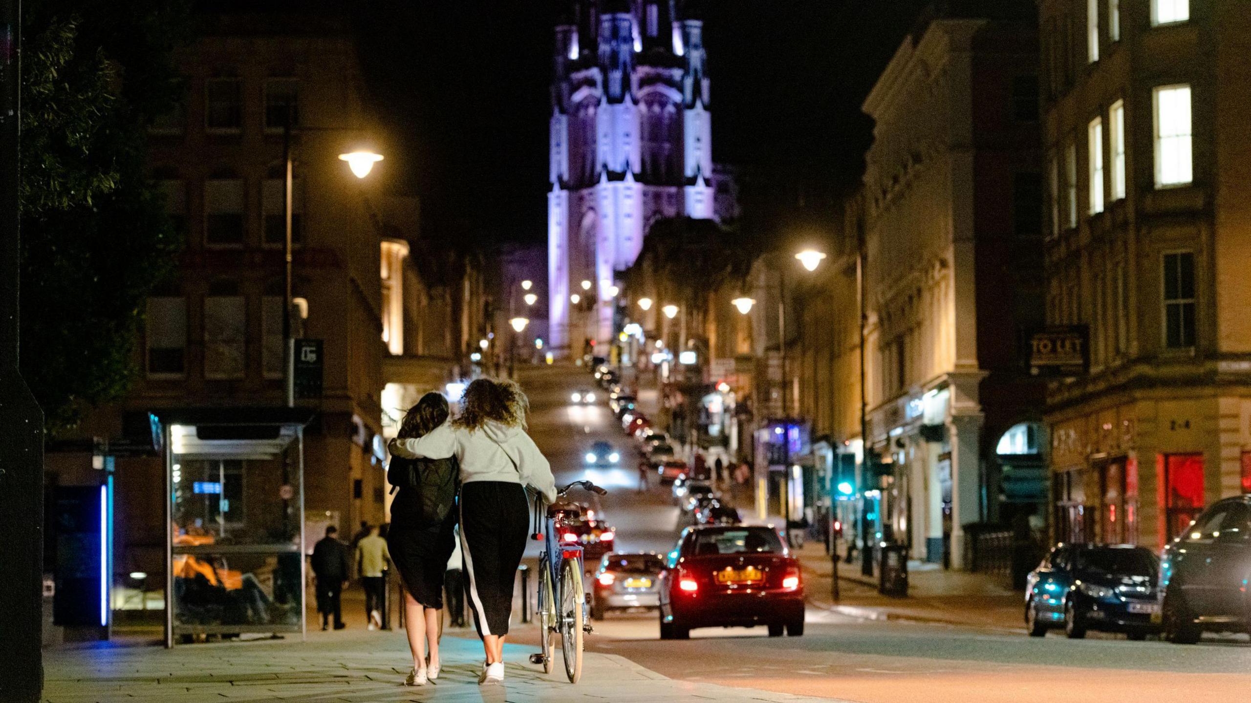 Two women are seen from behind walking towards Park Street in Bristol at night. They are both wearing black dresses and one, who is also pushing a bicycle, has her arm around the other. In the distance the Wills Memorial Building can be seen, lit up in purple light. Cars can also be seen on Park Street, driving in both directions with their headlights on