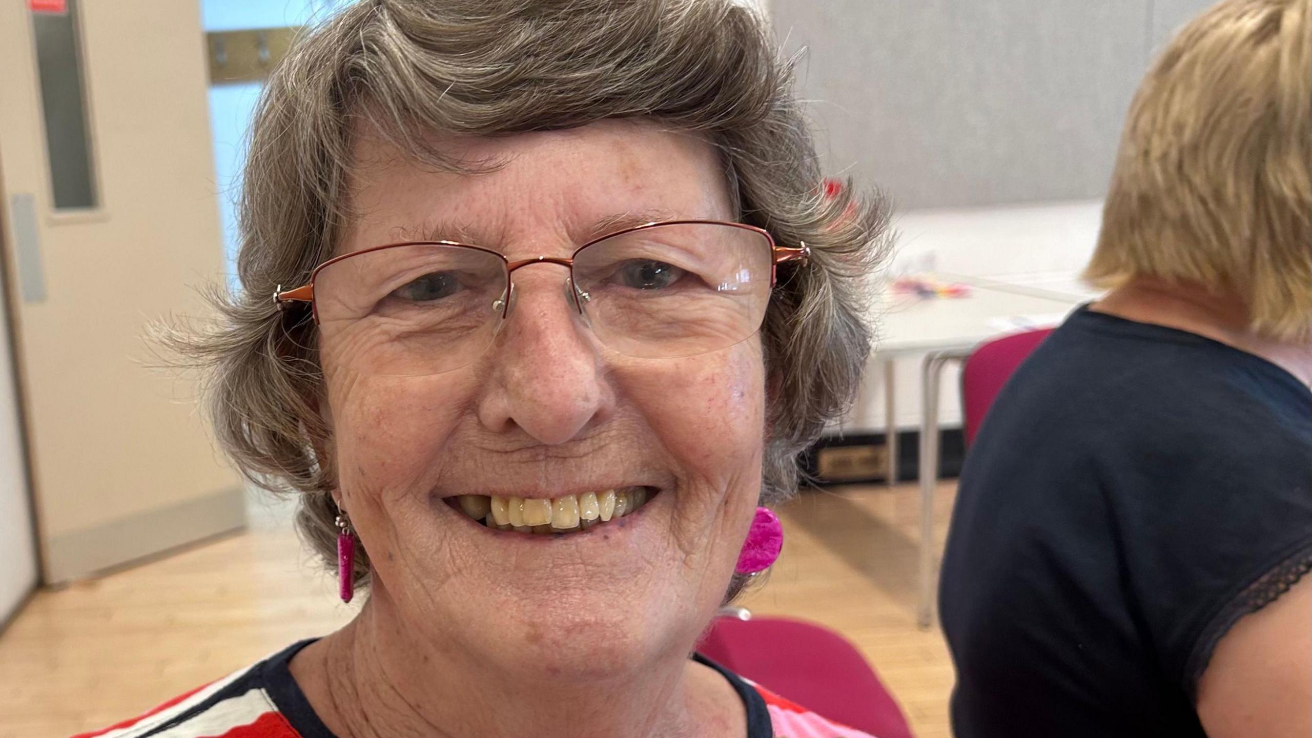 Norma Randall smiling at the camera in the village hall. She has pink earrings and rose gold-framed glasses