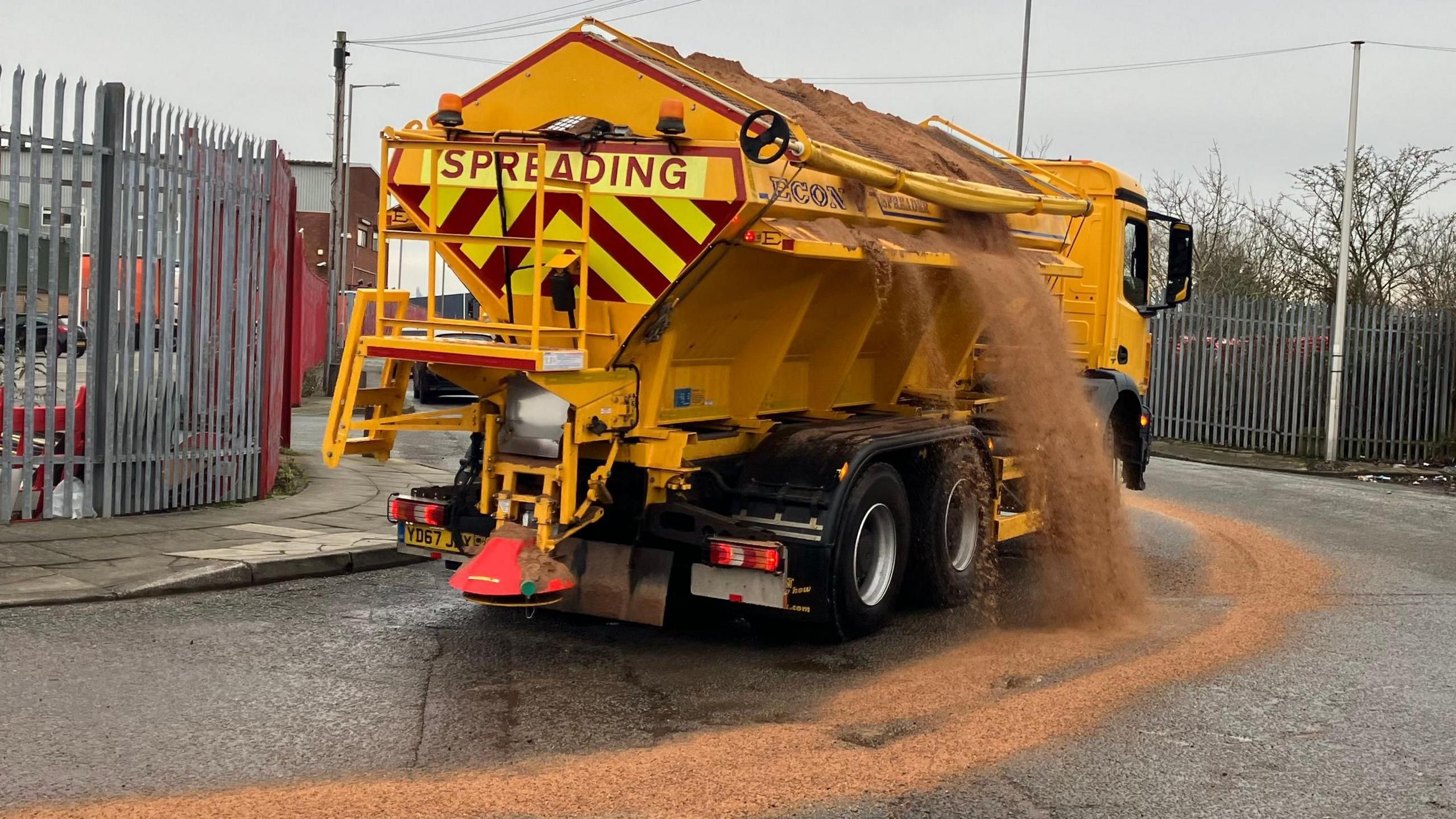 Yellow gritter dropping salt over road as it rides out of depot = SPREADING is in red letters on a yellow and red upside down triangle warning