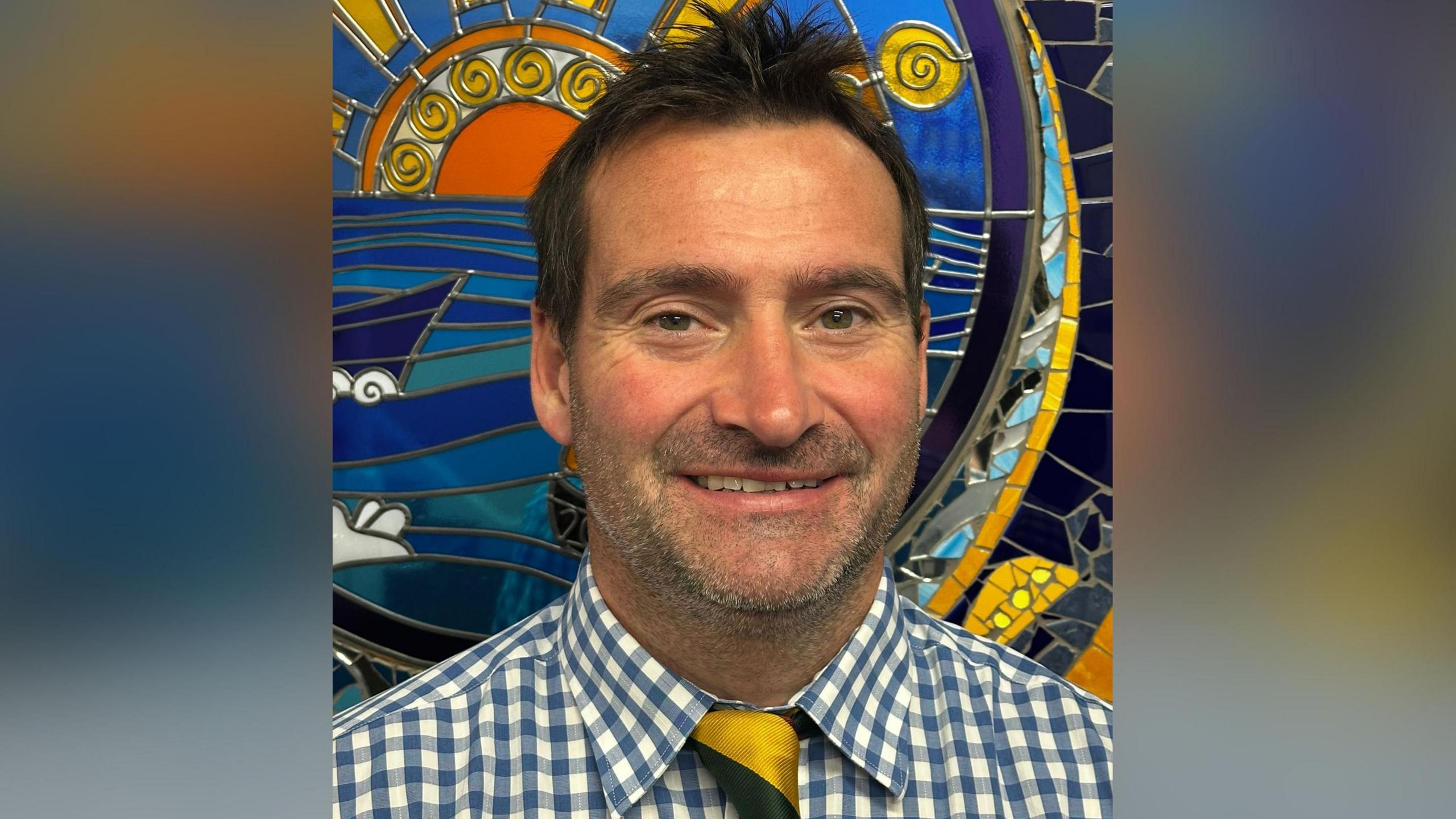 Llun o Tom Lewis yn gwenu ar y camera. Mae'n sefyll o flaen murlun lliwgar. Mae ganddo wallt a llygaid brown. Mae'n gwisgo crys siec glas a gwyn a thei melyn a du.