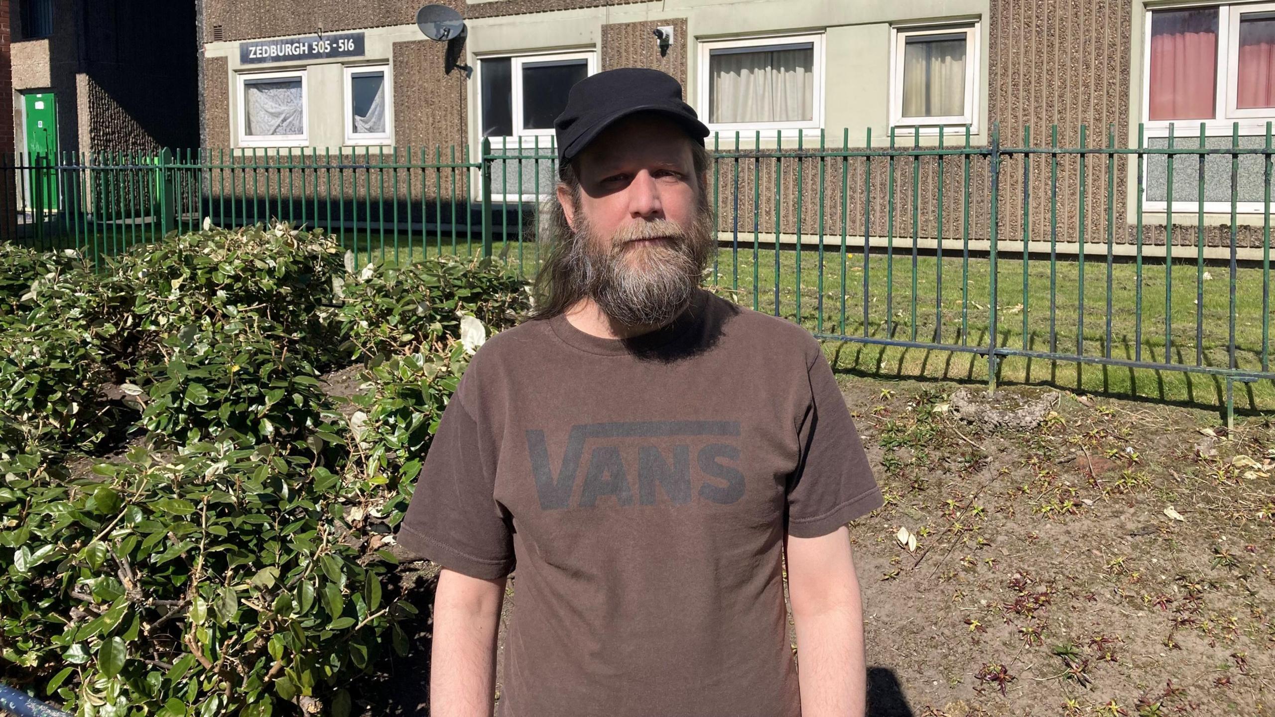 Andy stands in front of the flats. He wears a brown t-shirt with a black Vans logo and a black cap. He has a medium length brown and grey beard and long brown and grey hair,
