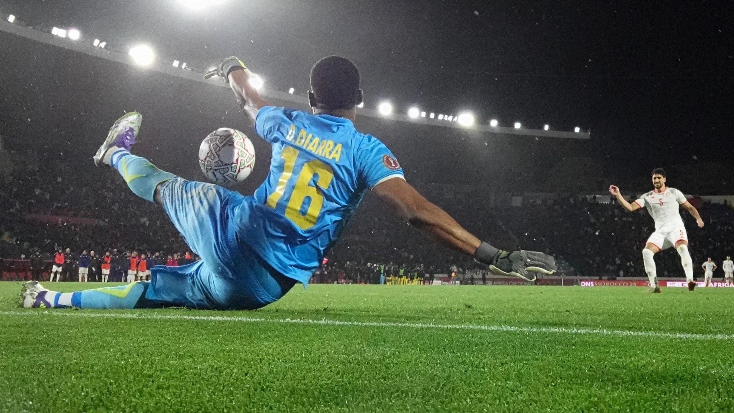 A picture taken from a ground-level angle inside a goal shows Mali goalkeeper Djigui Diarra saving a penalty from Tunisia's Mohamed Ali Ben Romdhane at the 2025 Africa Cup of Nations. Diarra, who is seen from behind, is in all-blue kit with his name and number 16 written in yellow. He is partially on the floor, with one leg touching the turf and the other raised, while his upper body is raised with arms spread wide. The ball is visible in the gap between his raised leg and one of his arms. Ben Romdhane can be seen in all-white an animated pose, having just kicked the ball. A large, full stand of the Casablanca stadium can be seen in the background, with bright floodlights on the top
