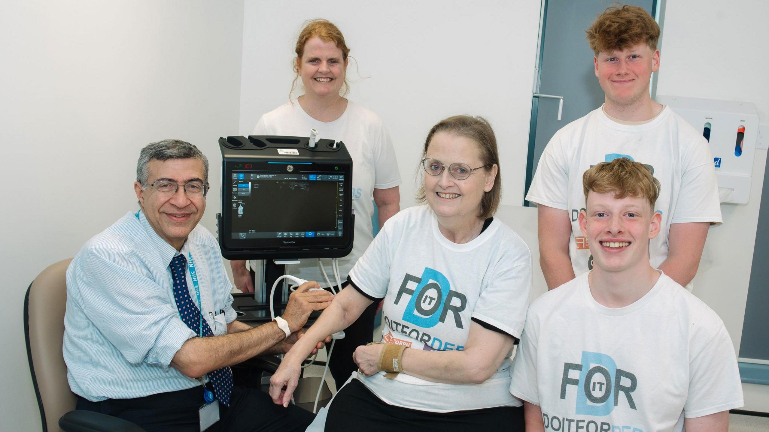 Dr Ayman Askari next to a black scanning machine. He is holding up a scanner to Debs Venezia, who is sitting to his right. Standing behind them is Rachael Griffiths, who has ginger hair. To their right, standing up, is Will Venezia and kneeling below him is Alex Venezia. They are all wearing which tshirts, which read "Do it for Deb"