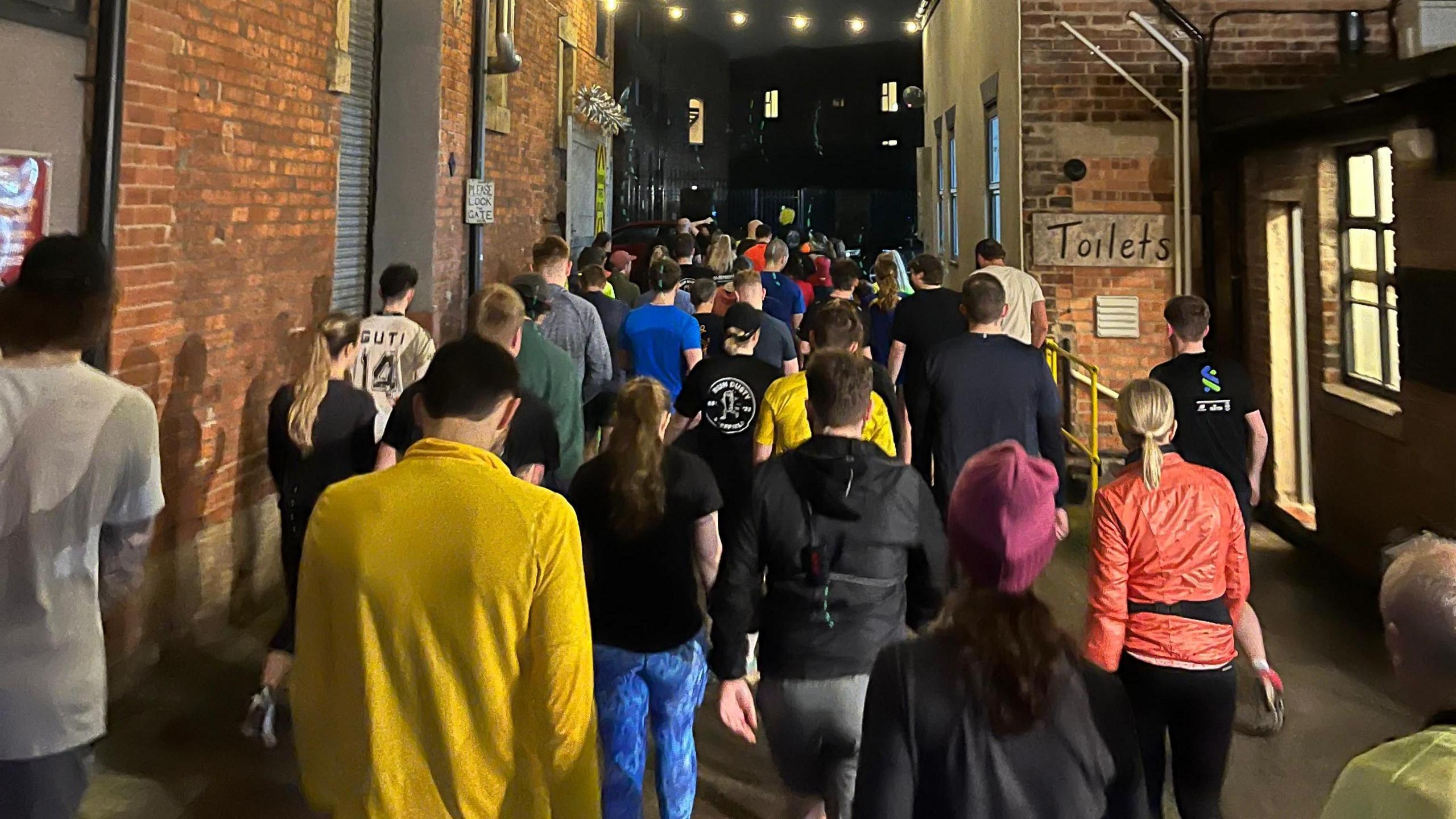 A group of people walking or jogging down a narrow alleyway lined with old brick buildings at night. Overhead, strings of warm lights are hung across the alley. The participants are dressed in athletic or casual clothing, with some wearing bright colours like yellow, orange, and blue.