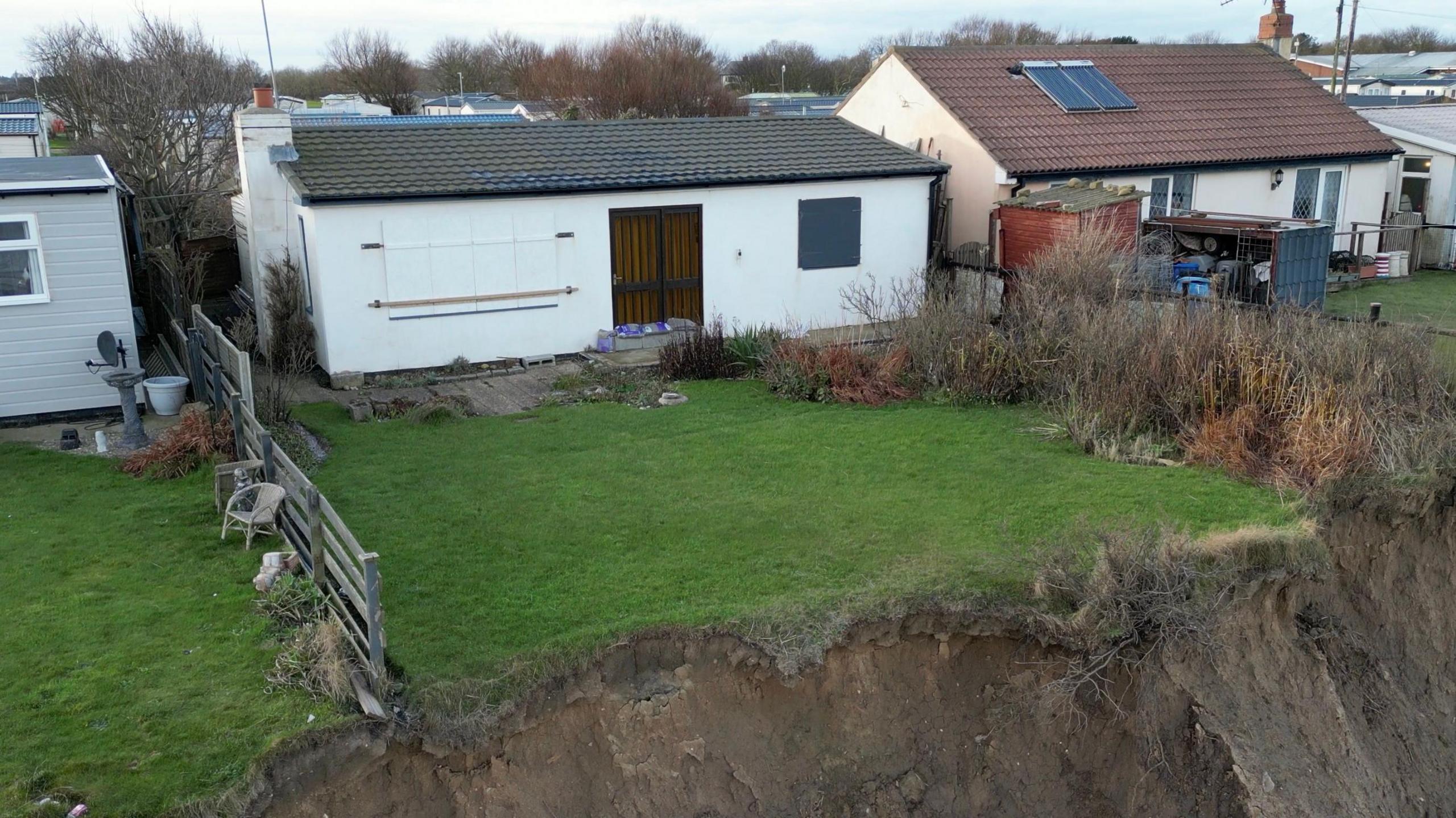 An exterior view of October Cottage in Skipsea. The property has been painted white and has grey roof tiles, a brown door and shutters over the windows. The property also has a grassed garden which is showing signs of erosion at the end which has become a vertical drop.