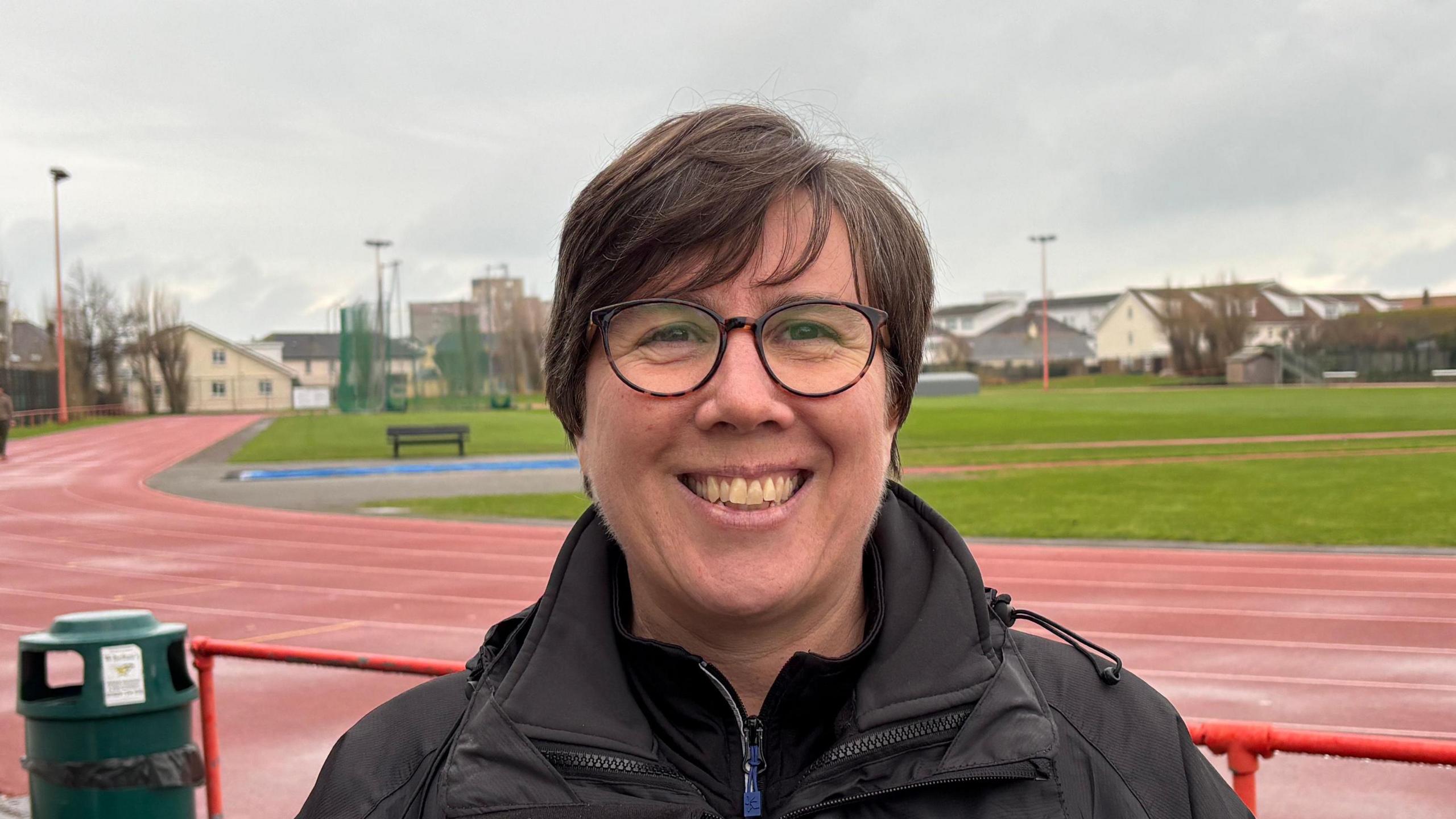 Rebecca Orpin, a woman with short, dark brown hair and brown rimmed glasses. She is wearing a black raincoat with a white badge. She is standing in front of a maroon athletics track, with a green field in its middle. She is staring directly at the camera and smiling.