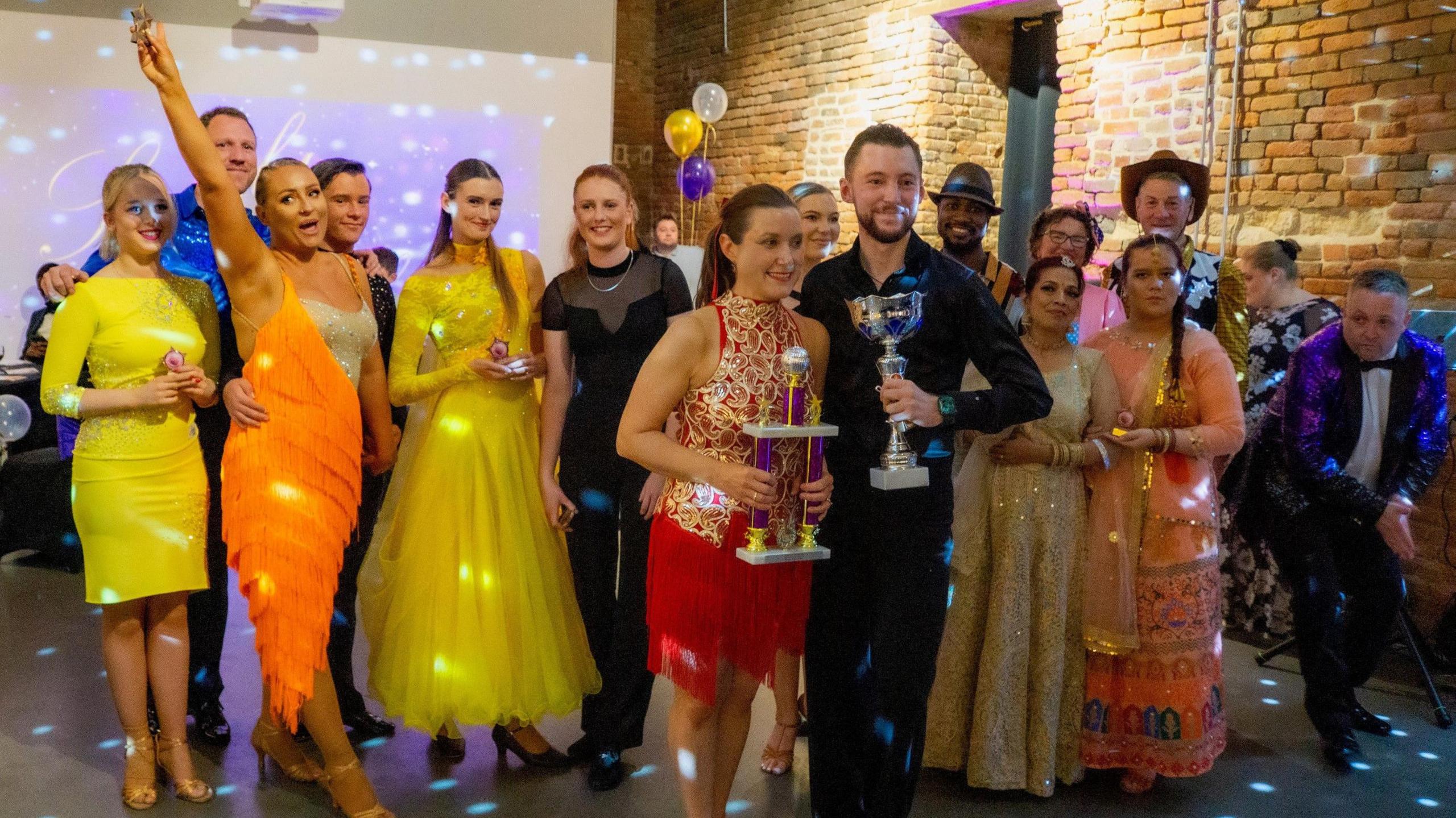 A group of people wearing ball gowns and evening wear in a party hall. A man and a woman in the front of the photo carry trophies in their hands