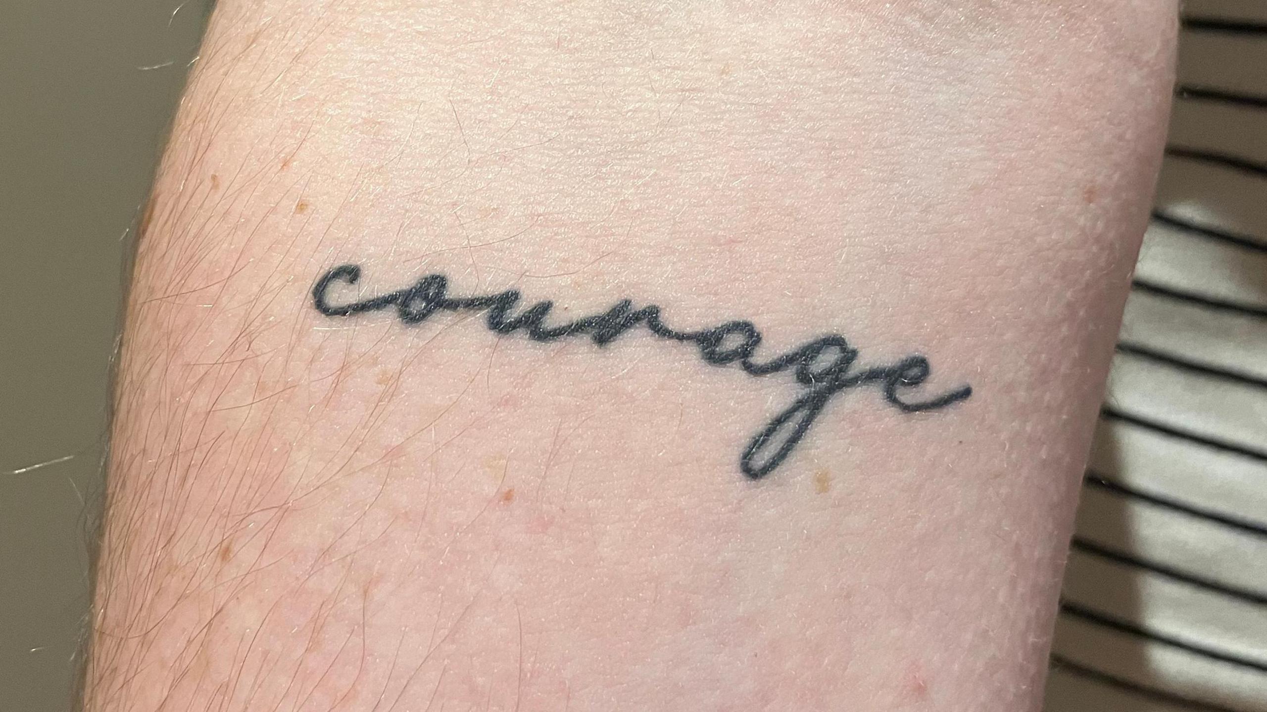 A woman is holding up her right arm.  She has a tattoo reading "courage" just below her elbow joint