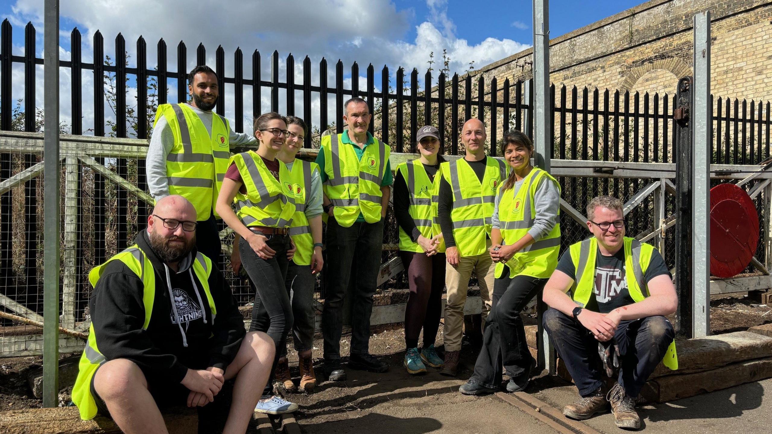 A group of volunteers wearing yellow, high visibility jackets are standing and sitting in front of former level crossing gates at the station