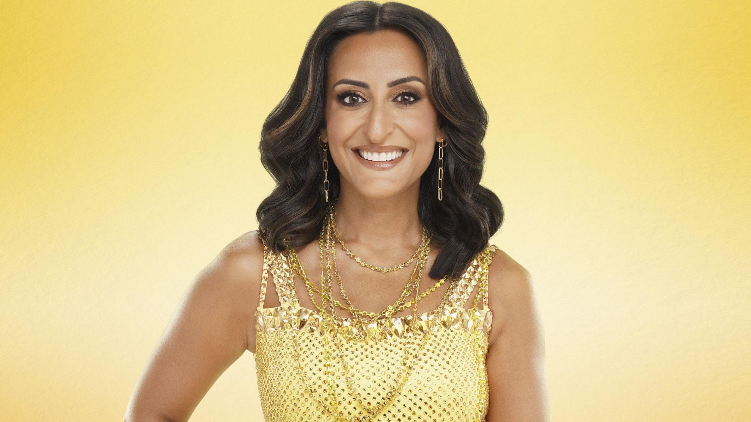 This image shows Strictly Come Dancing contestant Dr Punam Krishan wearing a yellow sequined dress and lots of gold jewellery. She has brown hair and gold eye makeup and is smiling at the camera. The photo has a solid yellow/gold background.