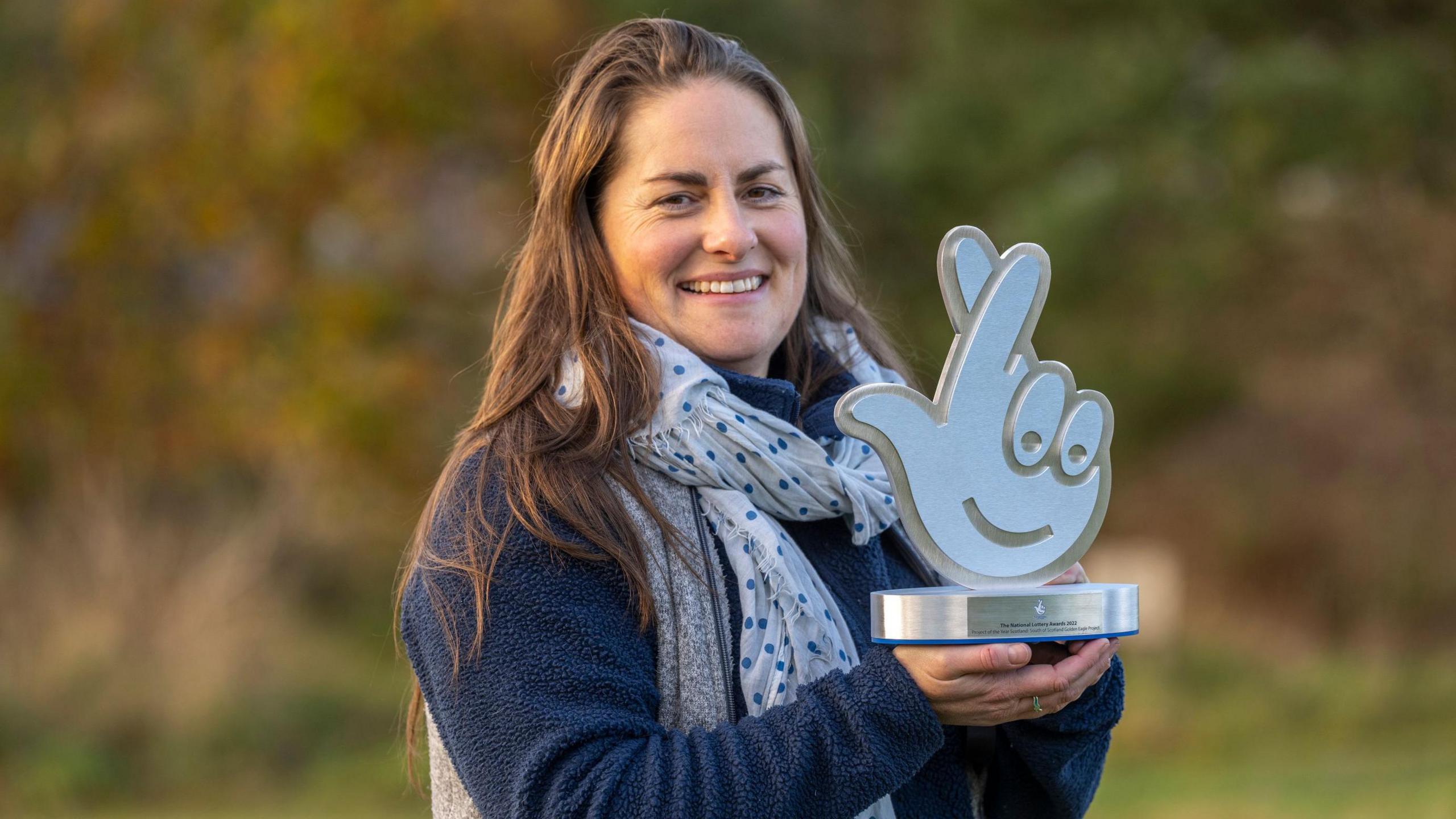 Dr Cat Barlow smiling as she holds up a trophy in front of a blurred countryside background