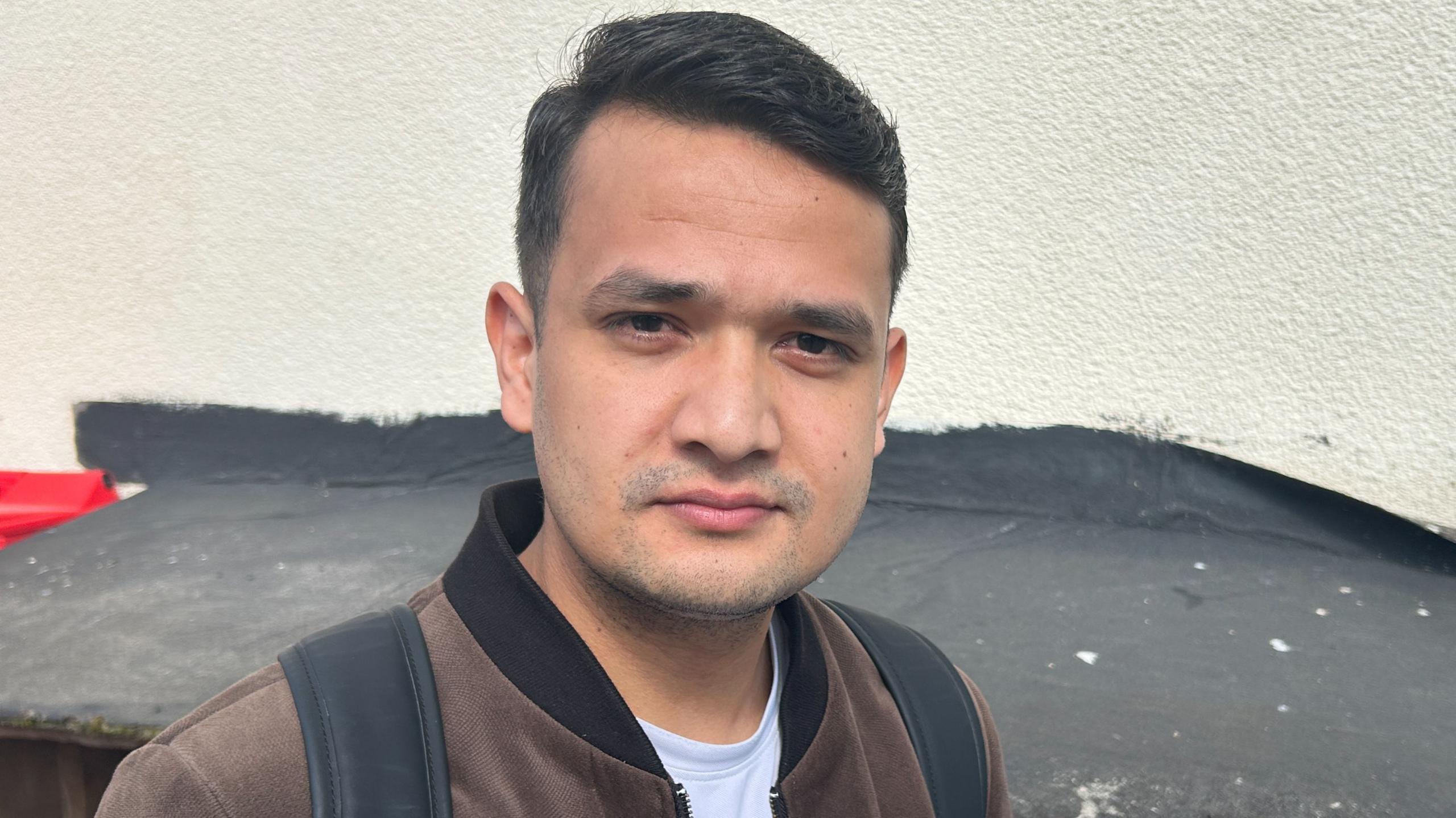 A man with dark brown hair combed in a side parting, wearing a brown velour bomber jacket over a white t-shirt, standing outside with a cream rendered building wall behind him.