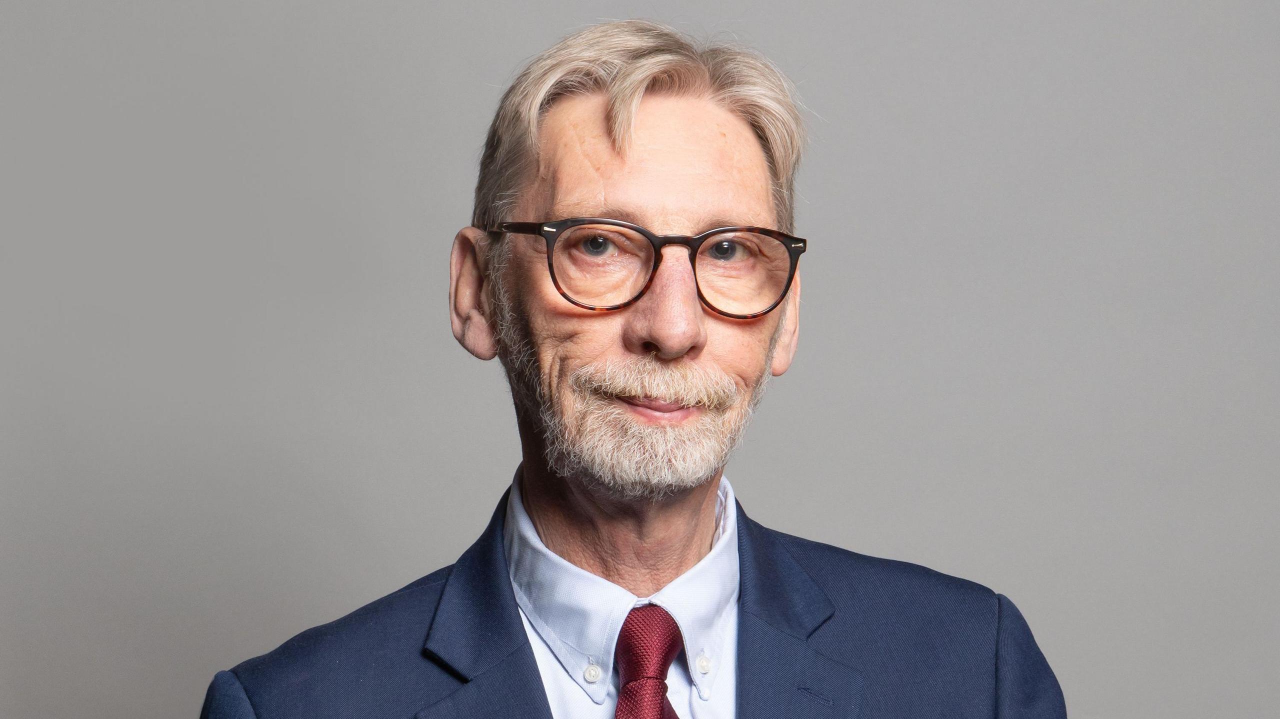 The official portrait of Sir George Howarth. He smiles into the camera, weating black glasses. He has gray, combed-over hair and a short beard. He wears a red tie. 