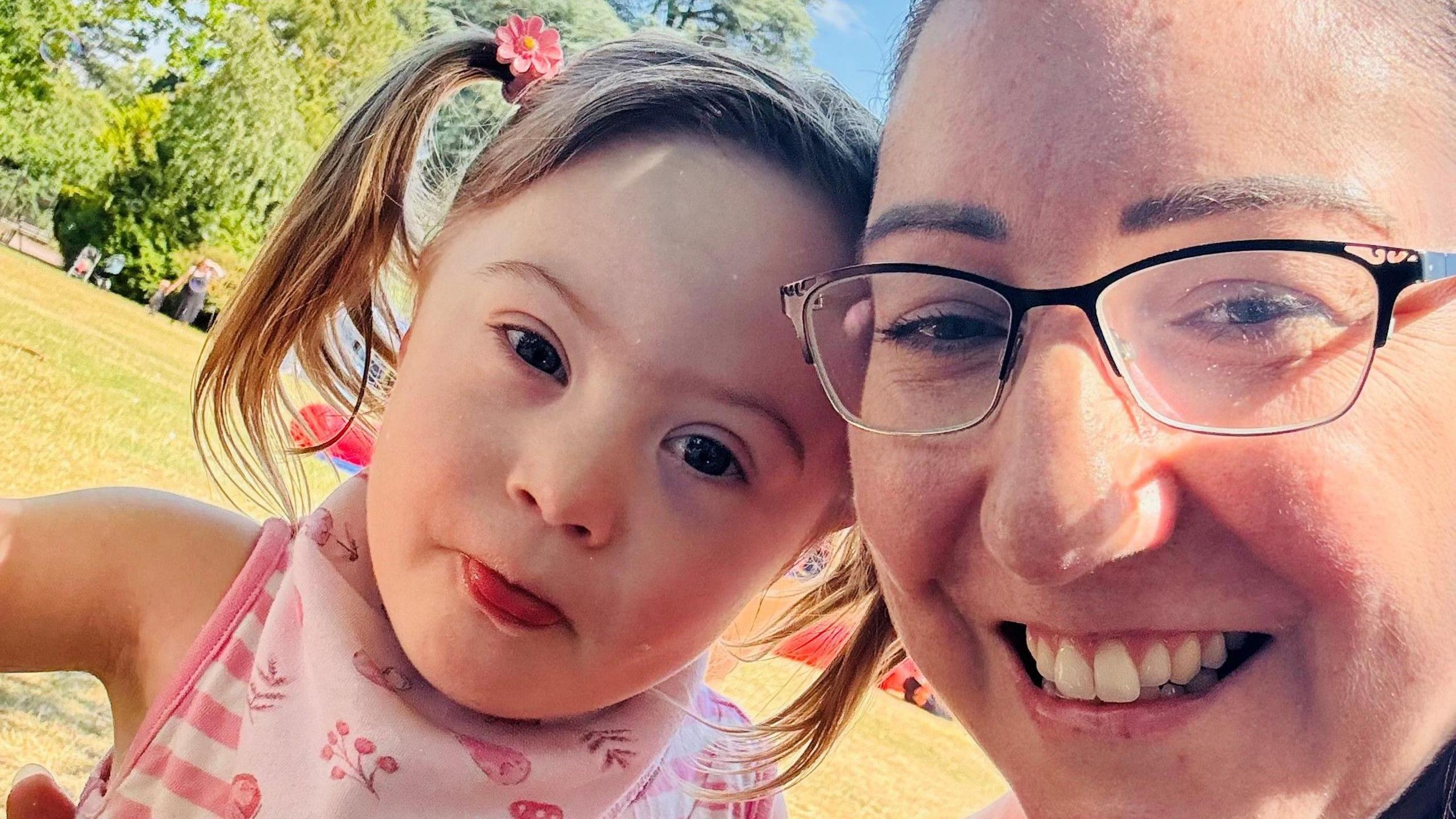 Violet and Rebecca smiling at the camera while sitting in a field at a park. Rebecca is wearing glasses with black rims. Violet is wearing a sleeveless pink top with her tongue poking out.