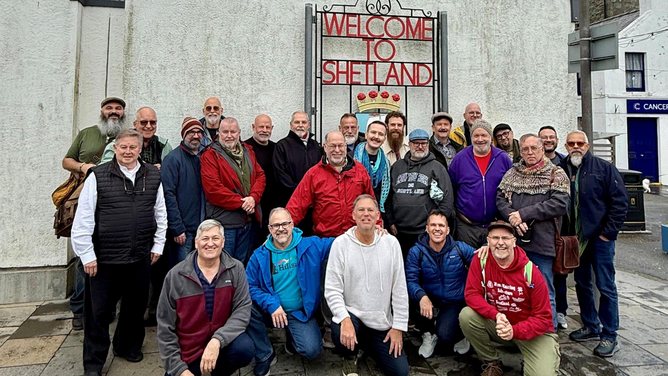 A group of men are standing/squatting in front of a sign that reads 'Welcome to Shetland'.