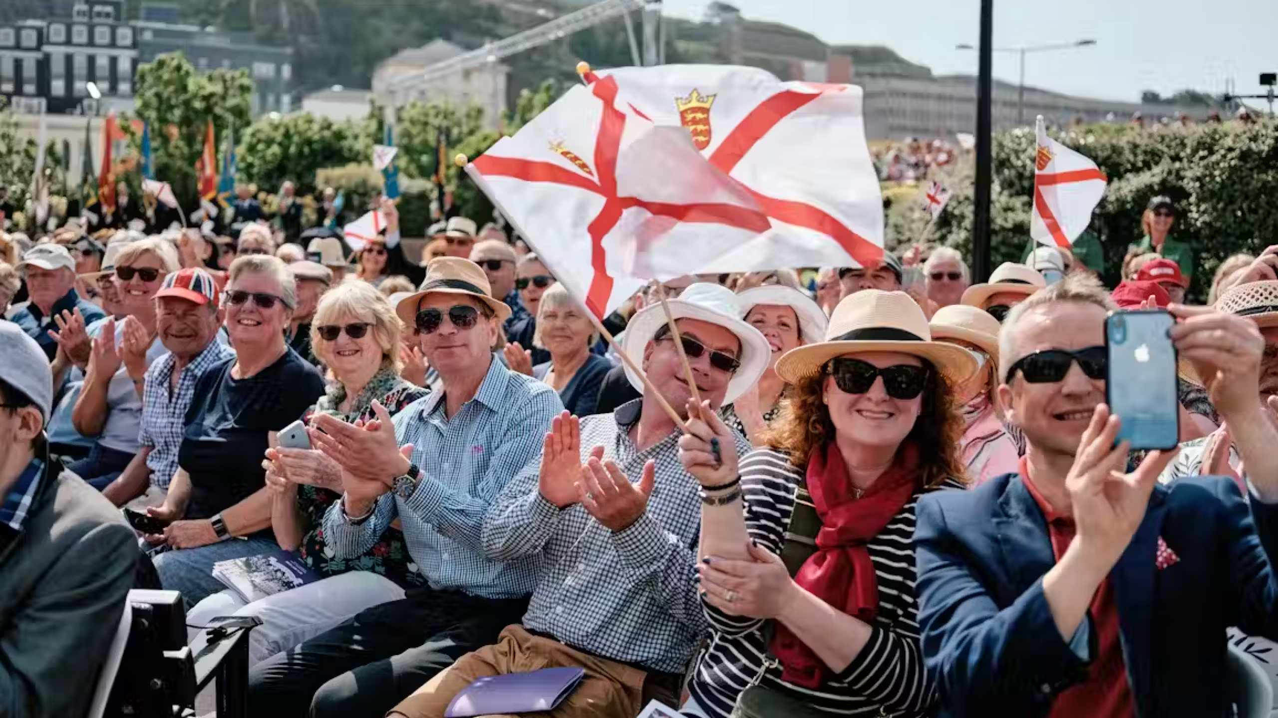 People are sat in a crowd and waving Jersey flags. They are clapping and smiling with a man on the right filming behind where the photo was taken. It is a large crowd with bushes behind with other properties.