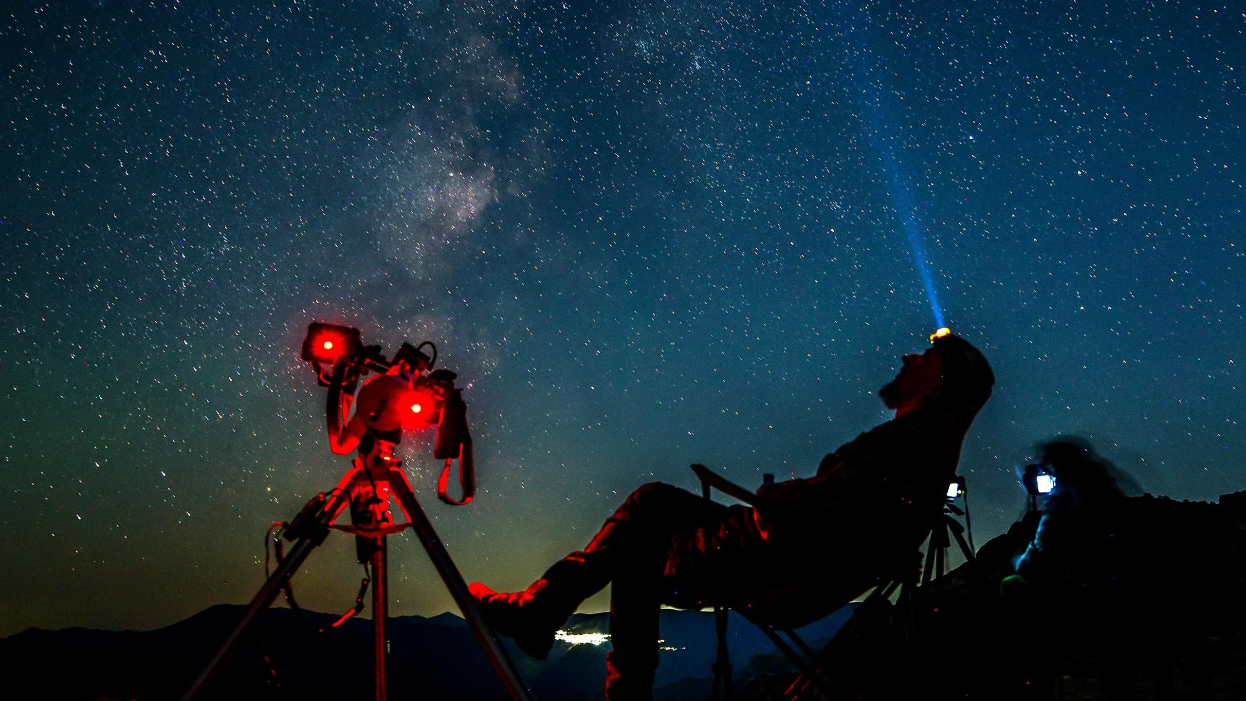 A man wearing a head torch sits next to a telescope at night