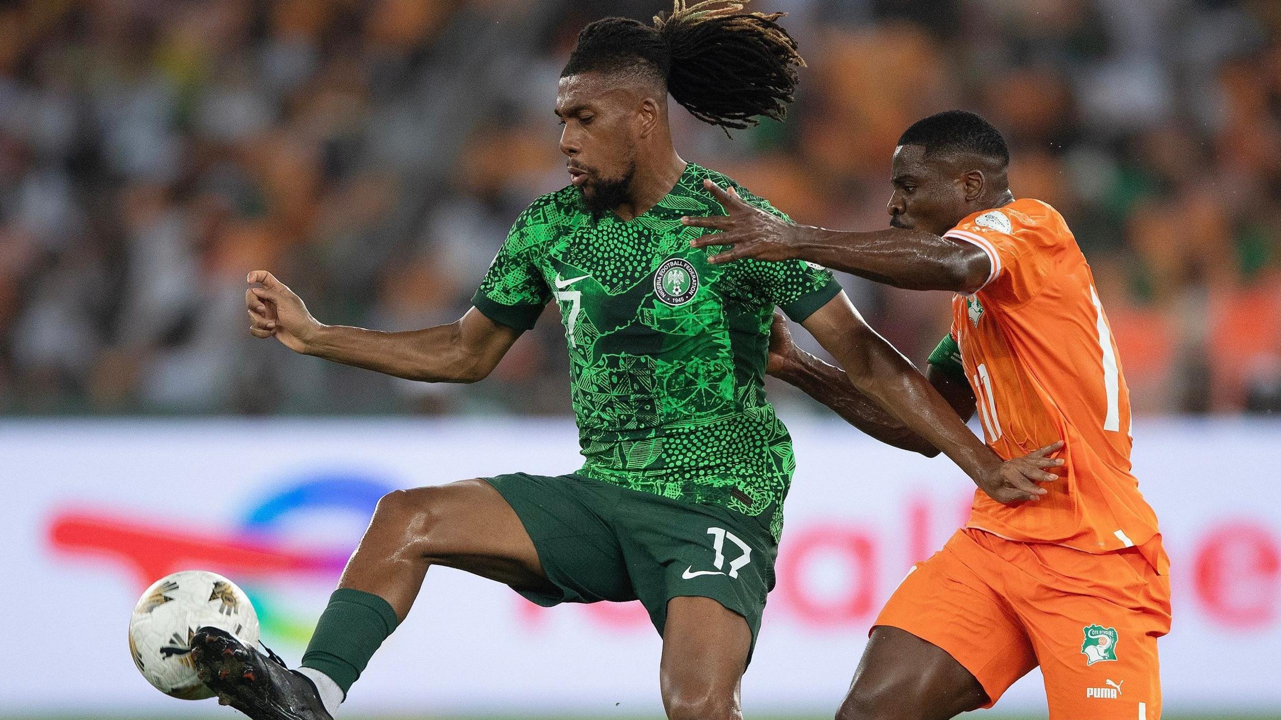 Alex Iwobi, wearing a green Nigeria kit, looks downwards as he controls a football with the outside of his right foot and holds off an Ivory Coast defender wearing orange with the palm of his left hand