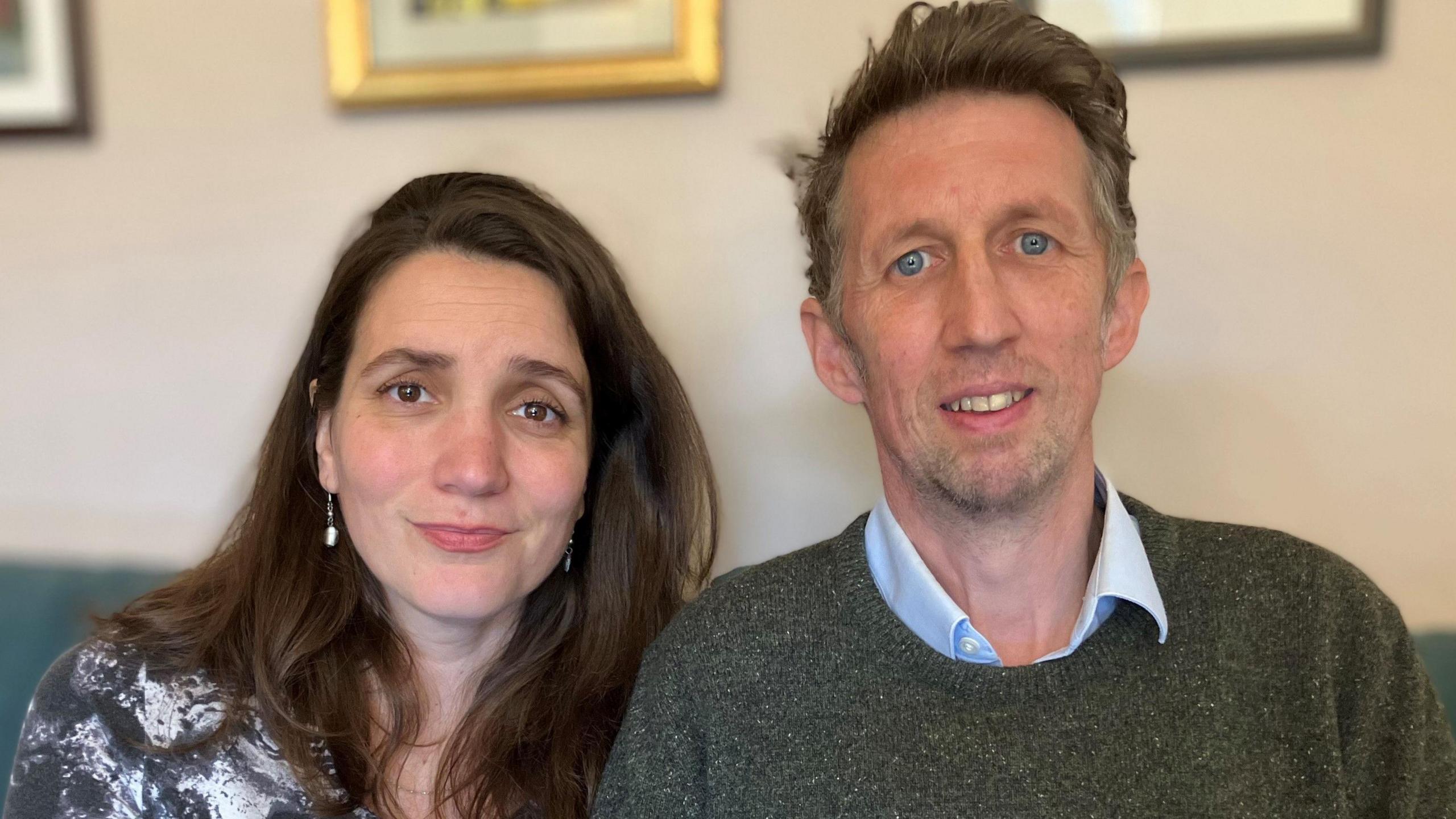 Andrew Davies (right) and his wife Milica. They are sitting on a green sofa and pictured from the chest up. She has long dark hair, is wearing a black and white marble-effect dress and drop pearl earrings. He has on a blue shirt with the collar unbuttoned and a dark green jumper over the top.