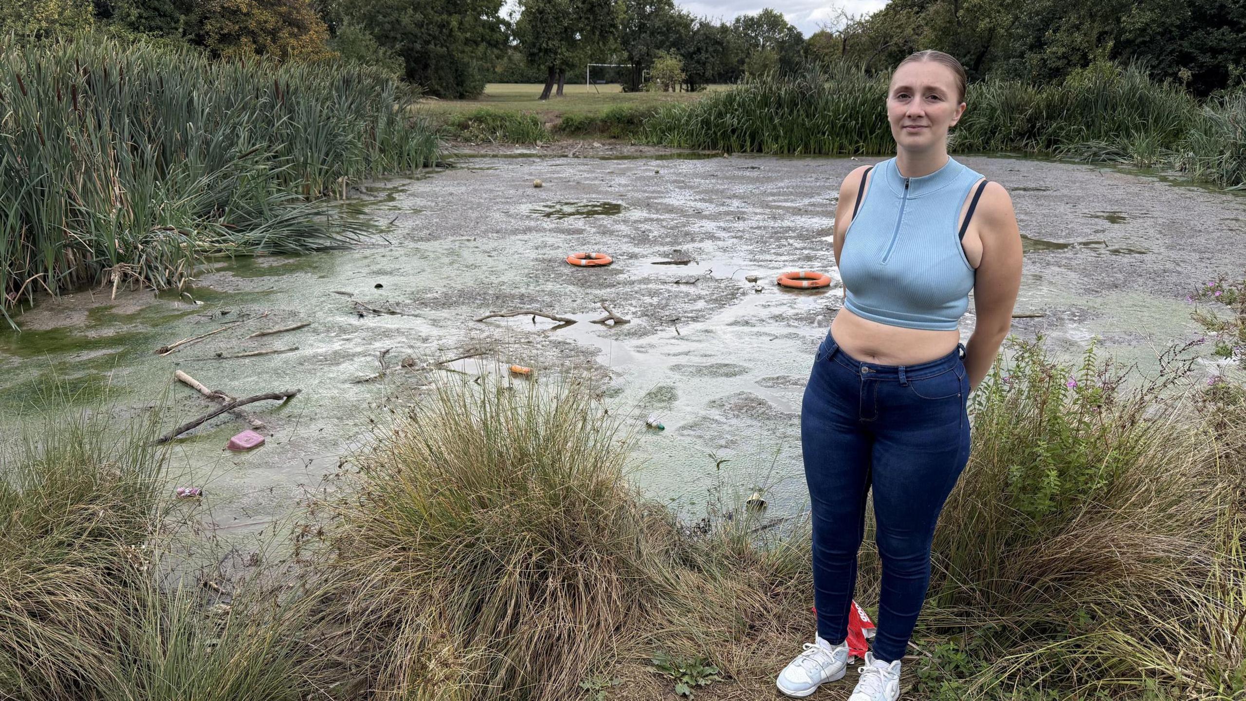 A woman in a blue gym top standing on the side of a lake that looks dark and unreflective due to the mud on top. Rubbish and two orange lifebuoys have been thrown into the water.