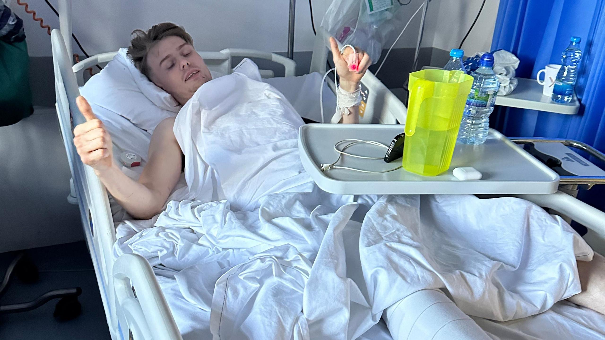 A young man in a hospital bed. He is laying down with his thumbs up in the air. His right leg is in plaster and there are drink bottles and a phone on a tray across the bed. He is in hospital.