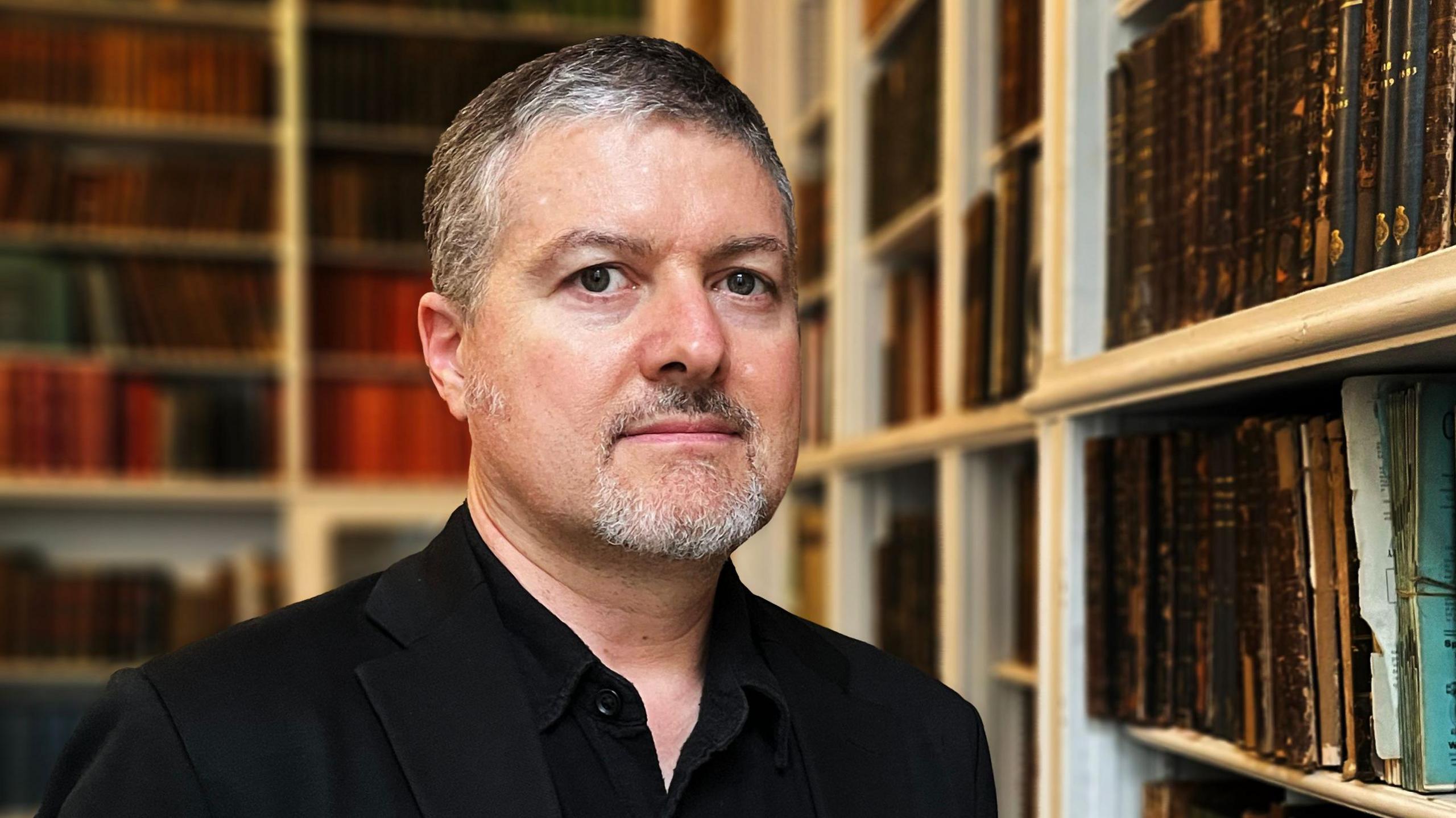 A man with grey-black hair and a beard is wearing a black shirt and standing in a room with shelves full of books.