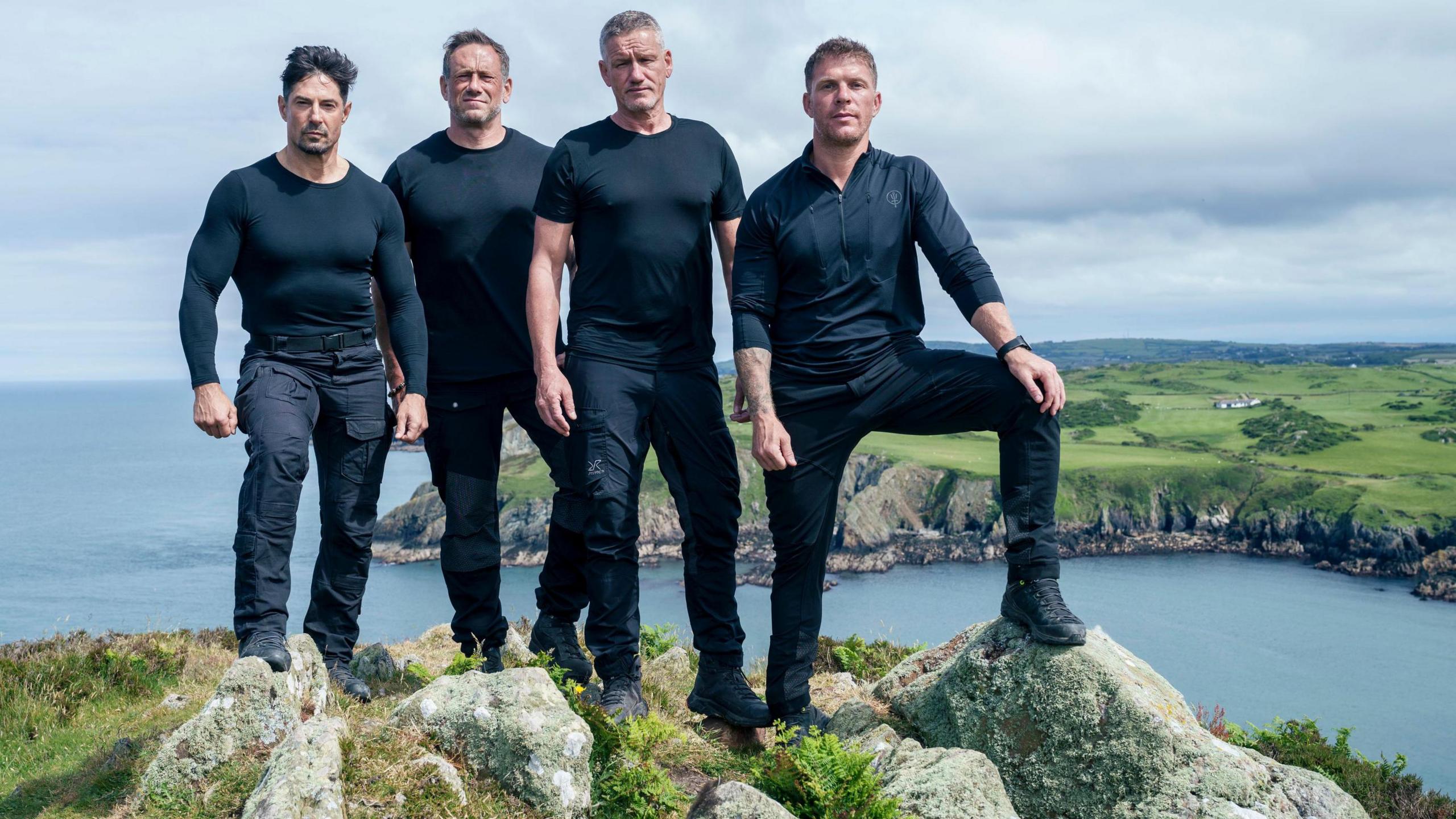 Four men in their black fatigues standing on a hilltop overlooking a coastline with a headland in the background and the sea beyond. The man on the right of the row perches his foot on a large rock.