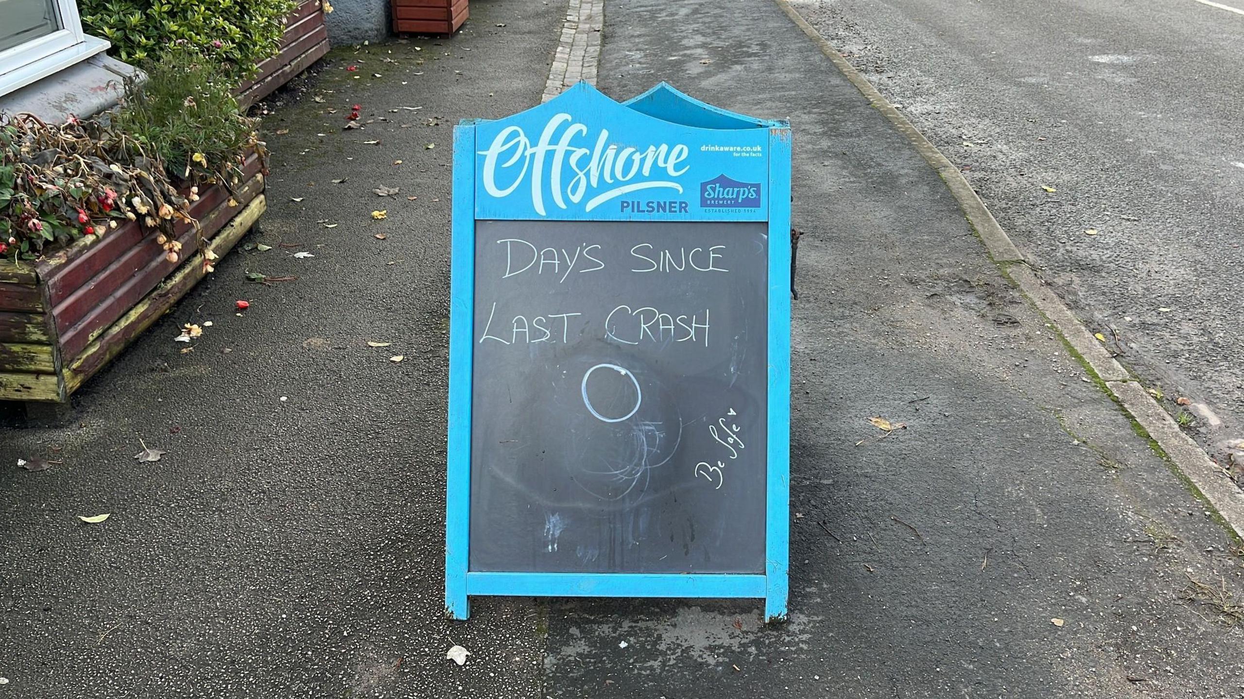 A blue-bordered A-board with a black chalkboard centre, with a message which reads "Days since last crash 0"