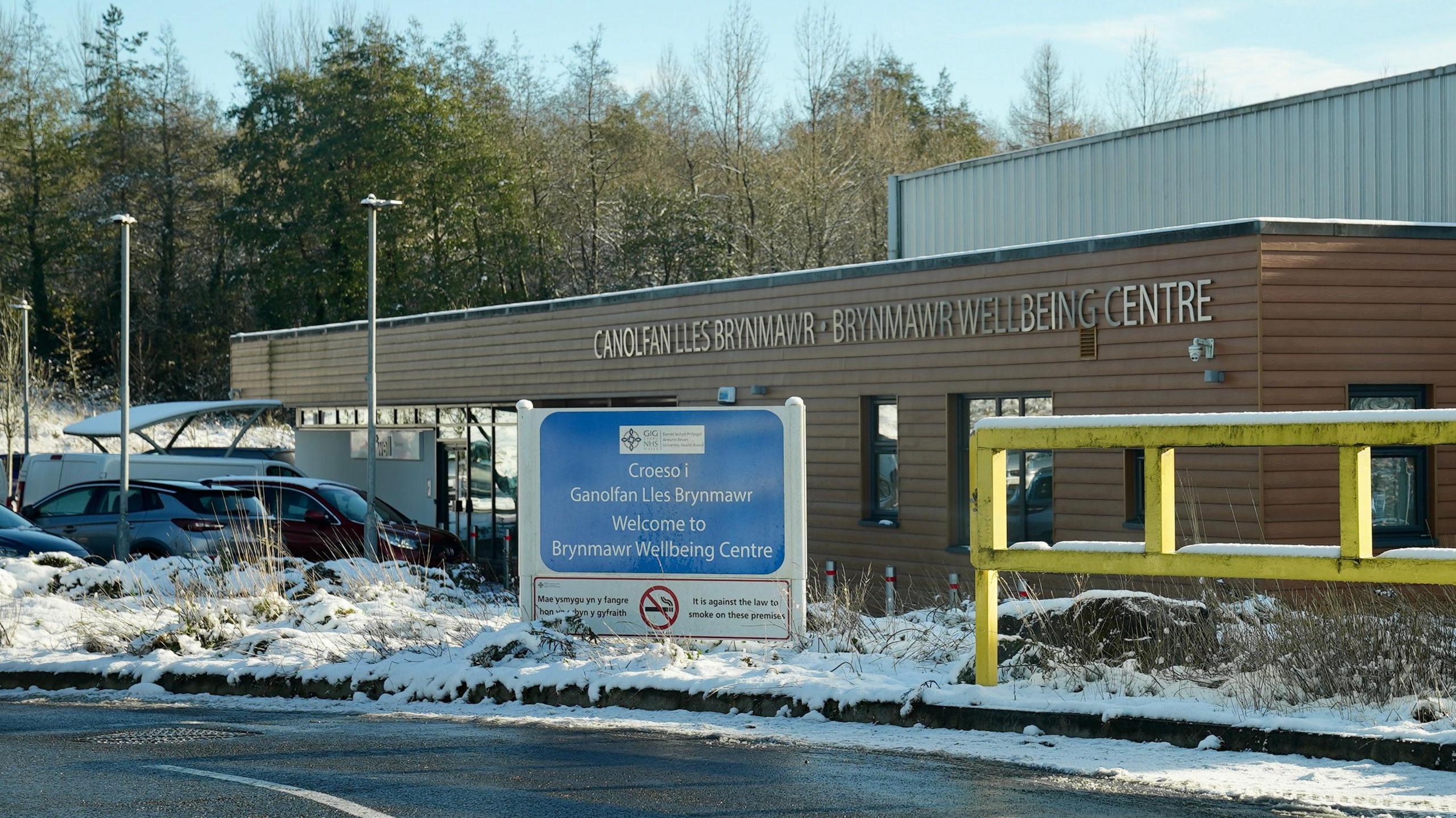 External view of the Brynmawr Wellbeing Centre, inside which Brynmawr Medical Practice is situated. It is a low building bearing its name in both English and Welsh, and there is a "Welcome" sign on the road nearby