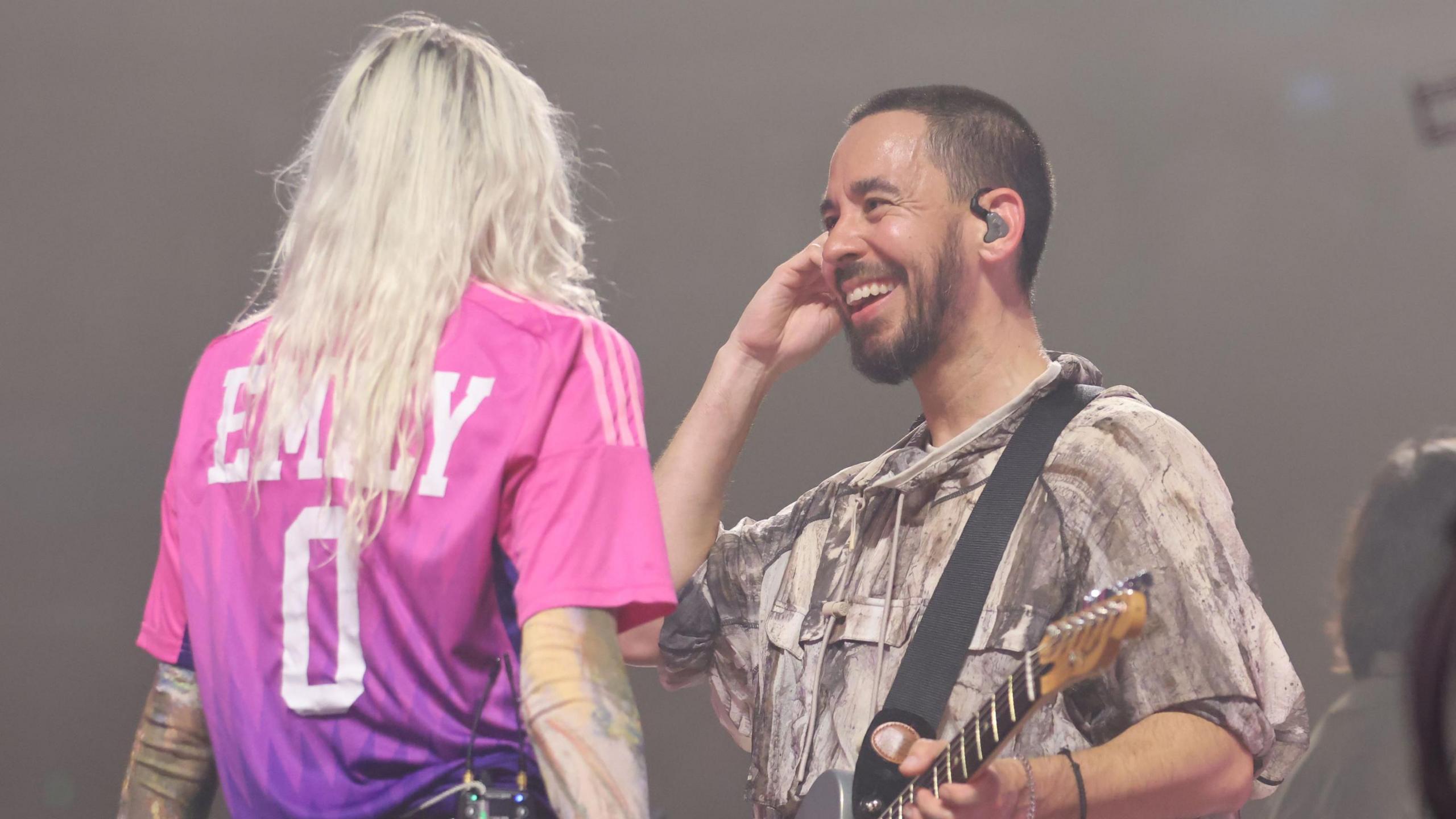 Emily Armstrong and Mike Shinoda on stage in Hamburg as part of Linkin Park's From Zero world tour. Mike smiles at Emily, who's facing away from the camera, her long bleached hair loose down her back as she wears a pink sports shirt with her name and '0' on the back. Mike has cropped dark hair and a short beard and holds a guitar in one hand and his earpiece in the other.