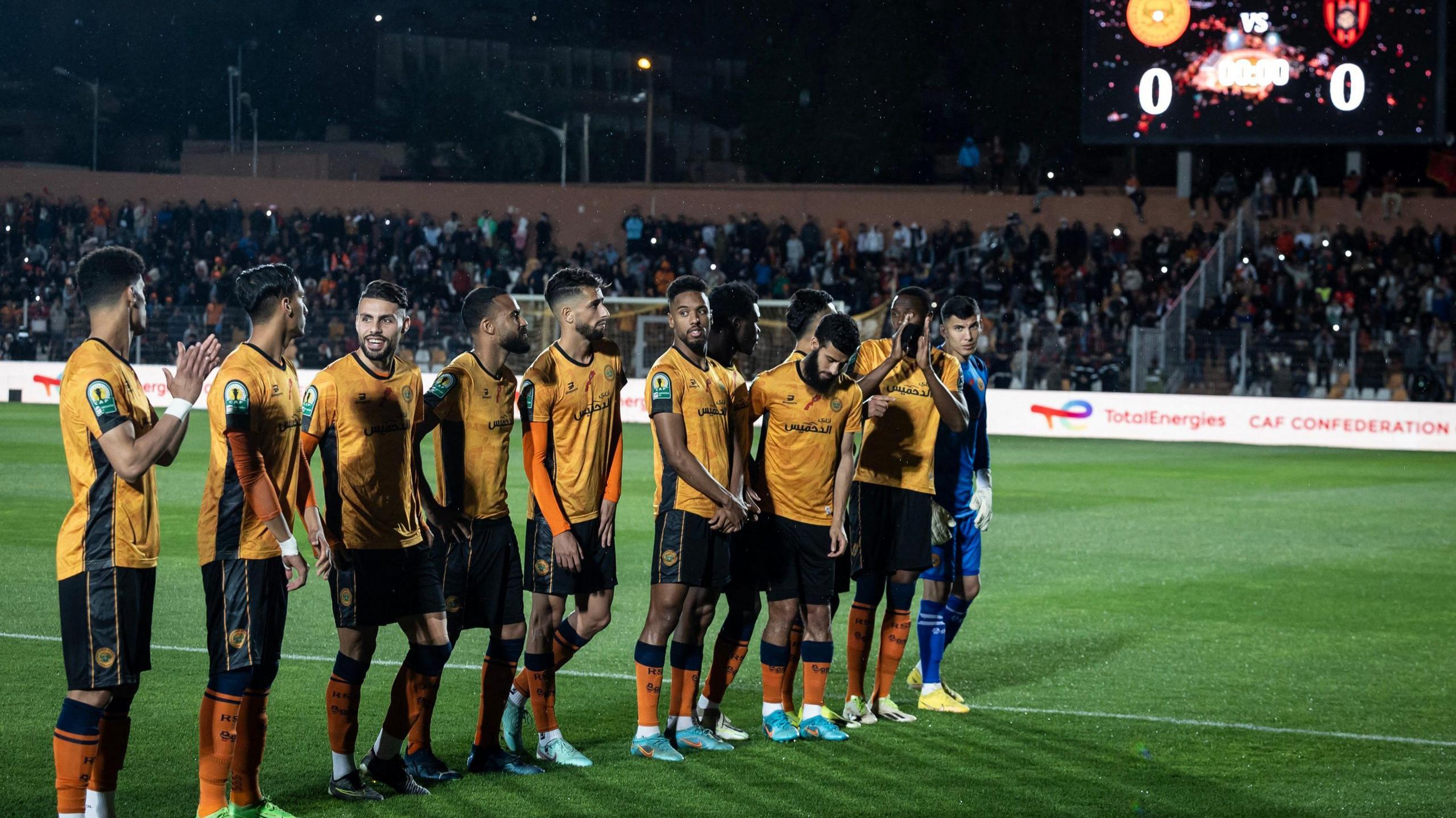 Eleven RS Berkane players, wearing orange shirts, black shorts and orange socks, line up on a pitch ahead of a game