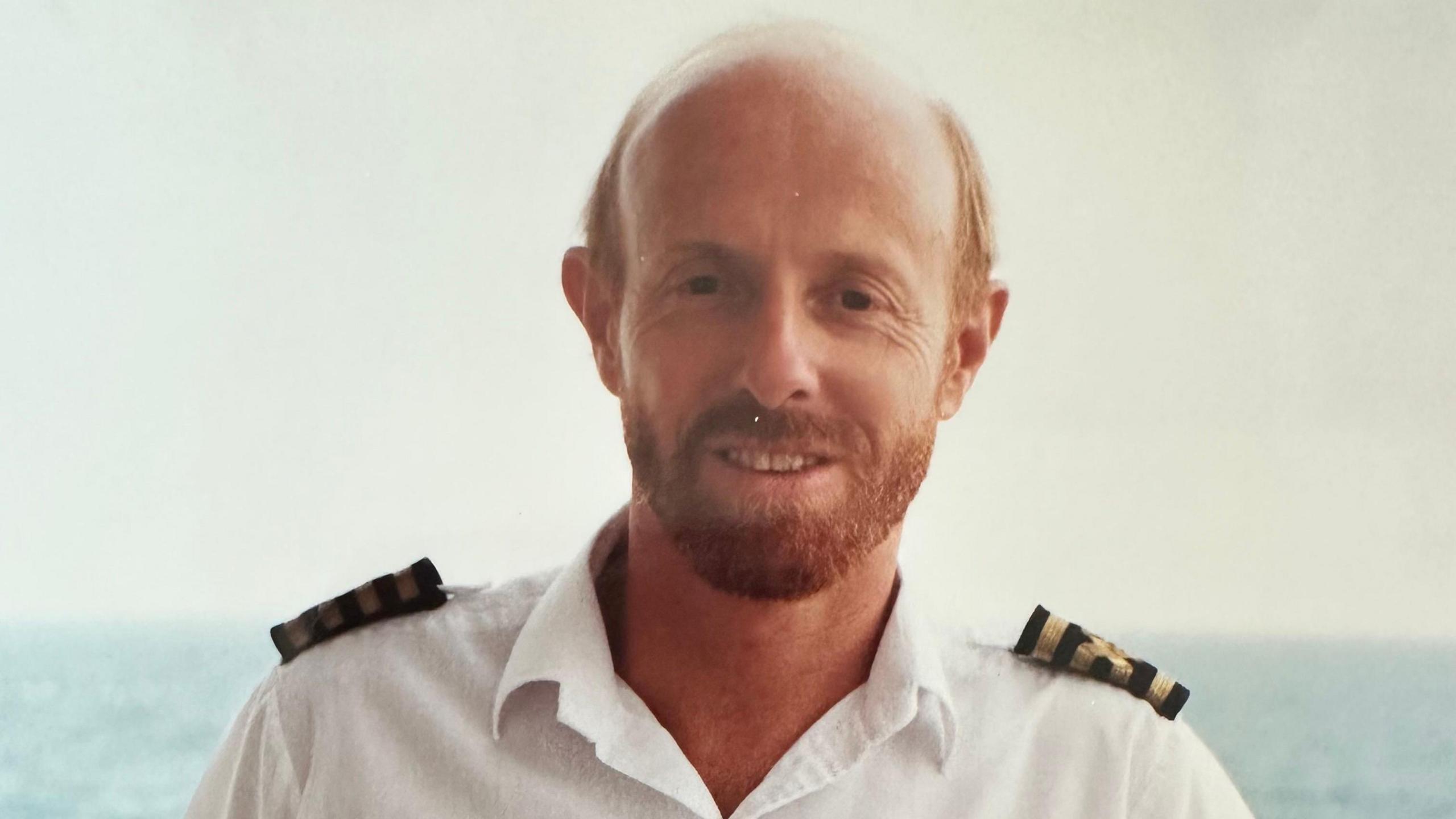 An old photo of Barry Dean smiling into the camera in his navy uniform, which is a white shirt with navy blue and gold shoulder patches. He has ginger hair and beard and the sea can be seen behind him.