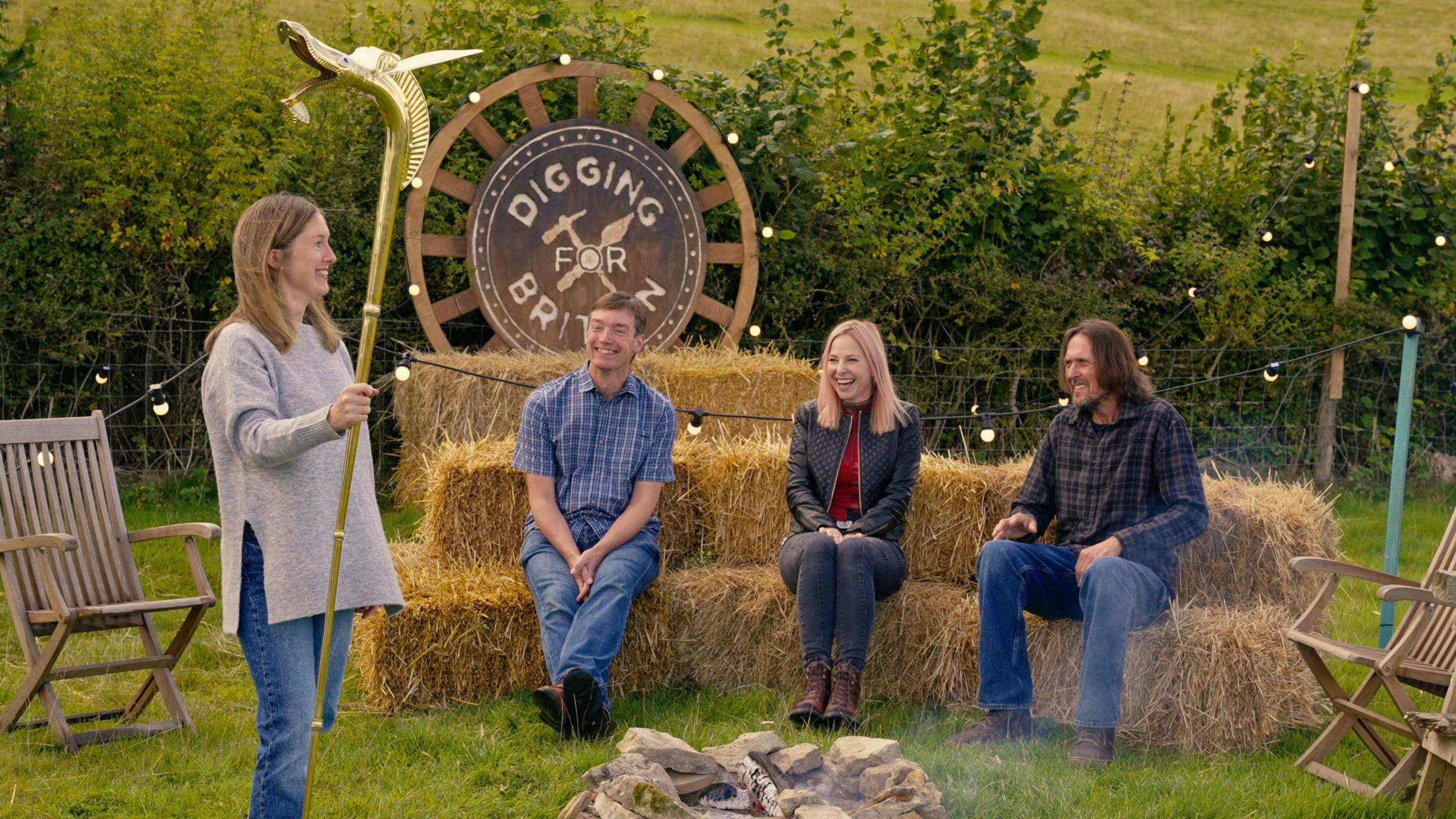 A scene in a field showing a group of people during the filming of Digging for Britain. A woman wearing a jumper and jeans is on the left, holding a brass coloured carnyx with a gaping mouth in her right hand. In the middle are straw bales and sitting on them is a man in a paid shirt and blue jeans, a woman in black jacket and black jeans and a man in plaid shit and blue jeans. They are all laughing. 