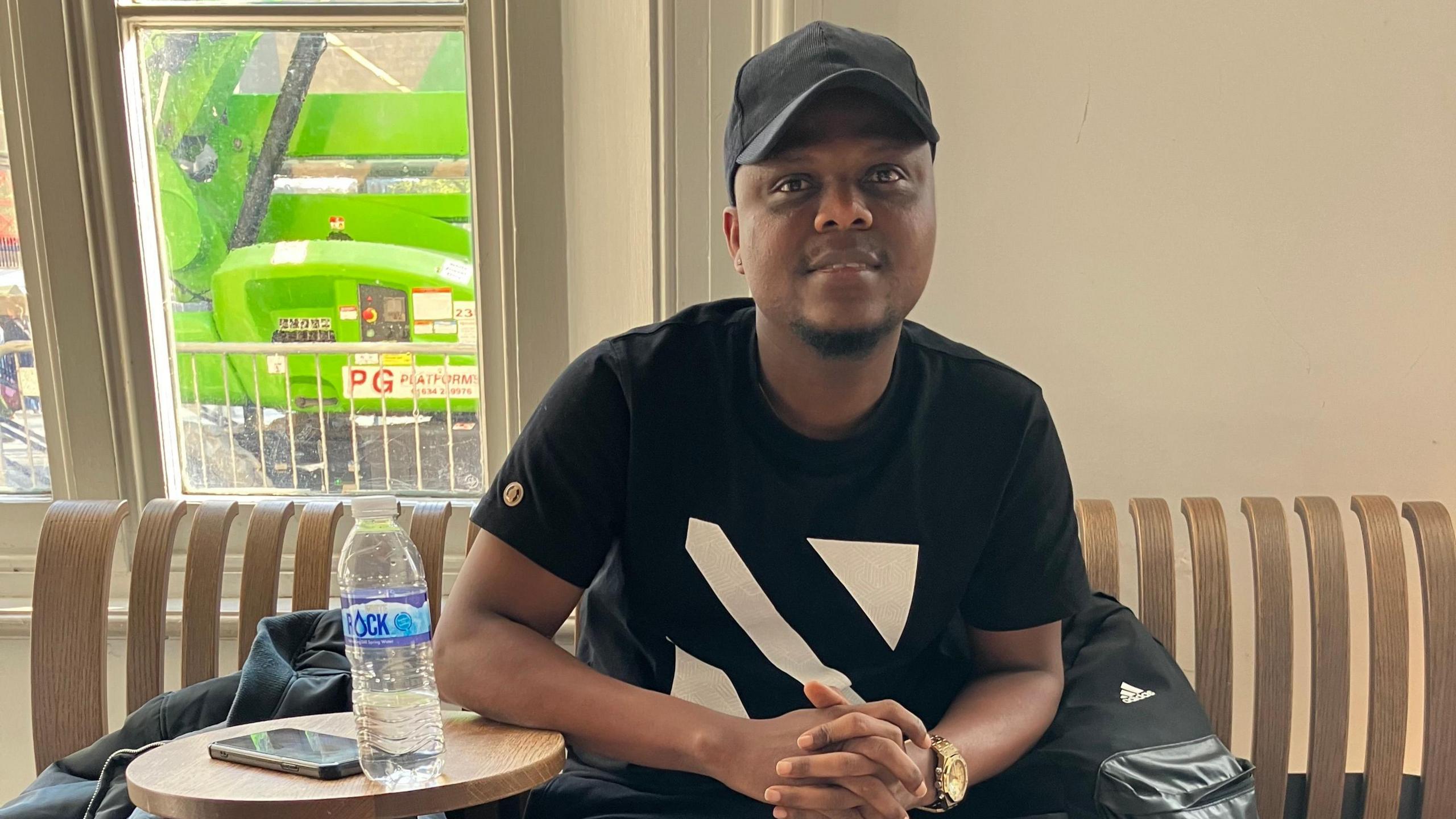 Israel Obinusi, a black man wearing a black hat, a black t-shirt and a gold watch, sits on a trendy slatted bench at Kings Cross station in London