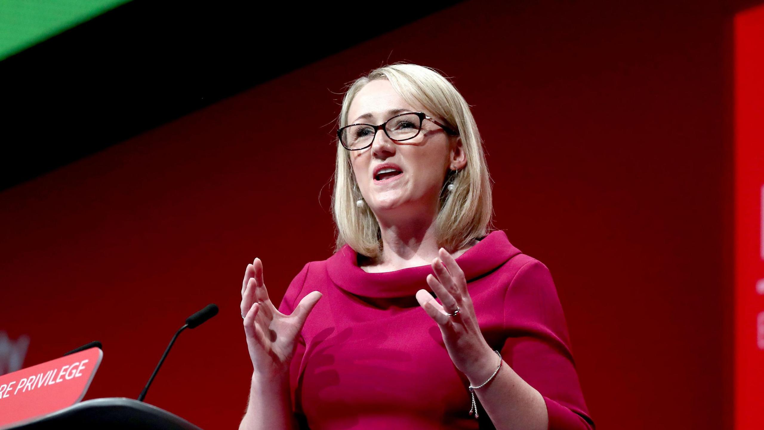 Rebecca Long-Bailey stands at a podium at a Labour party conference, delivering a speech.