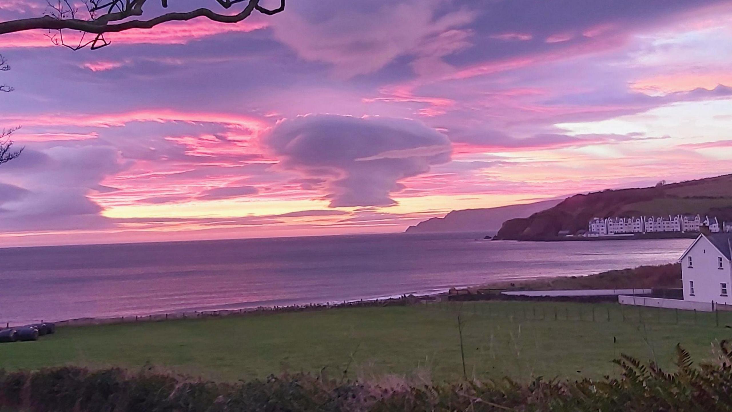 A cone shaped cloud has been photographed during sunrise. The sky is a mix of purples, pinks and yellow. In the foreground is a green field and a number of white houses, beyond it is the sea. 