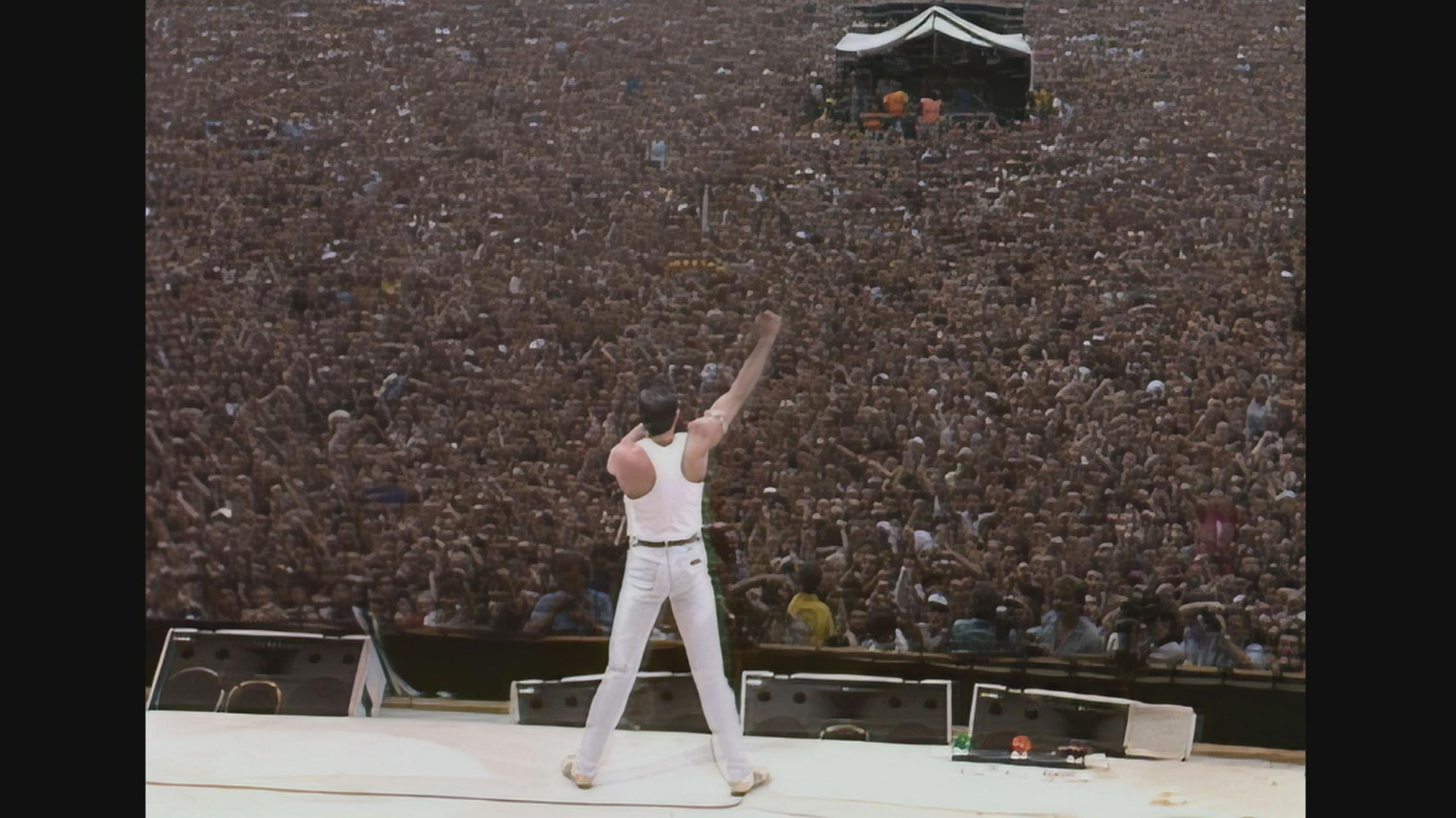 Freddie Mercury stands on the stage at Live Aid facing the packed audience. We see him from behind in a wide stance, he is dressed all in white - trainers, jeans, tank top. He holds the microphone to his mouth and his right arm is raised in a fist. 