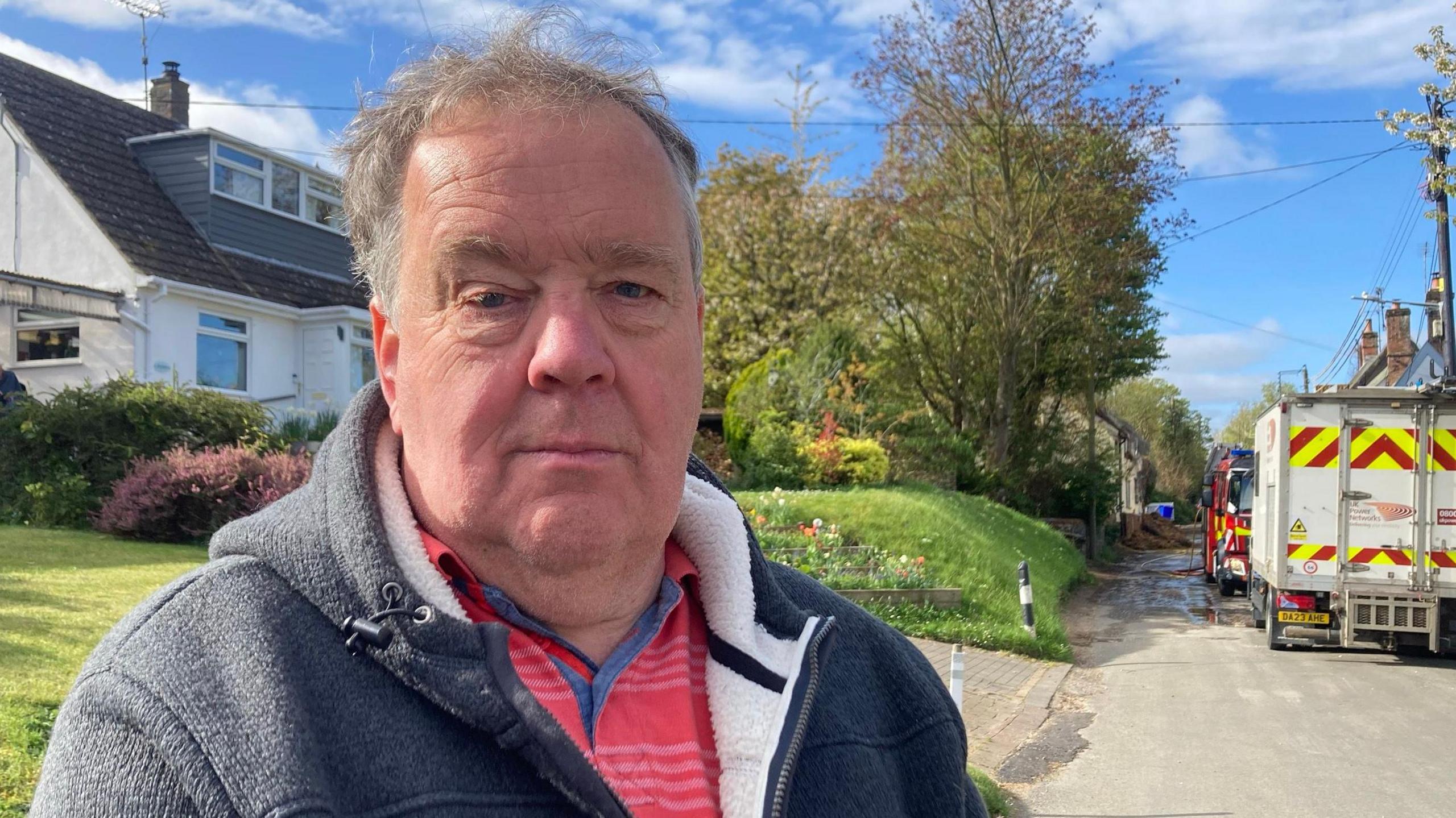 David Ruffles is standing on the left looking at the camera. He has short grey hair, and is wearing a red and white stripped shirt, and a grey zip up hoodie. On the road where he is standing there is a fire engine parked in the middle.