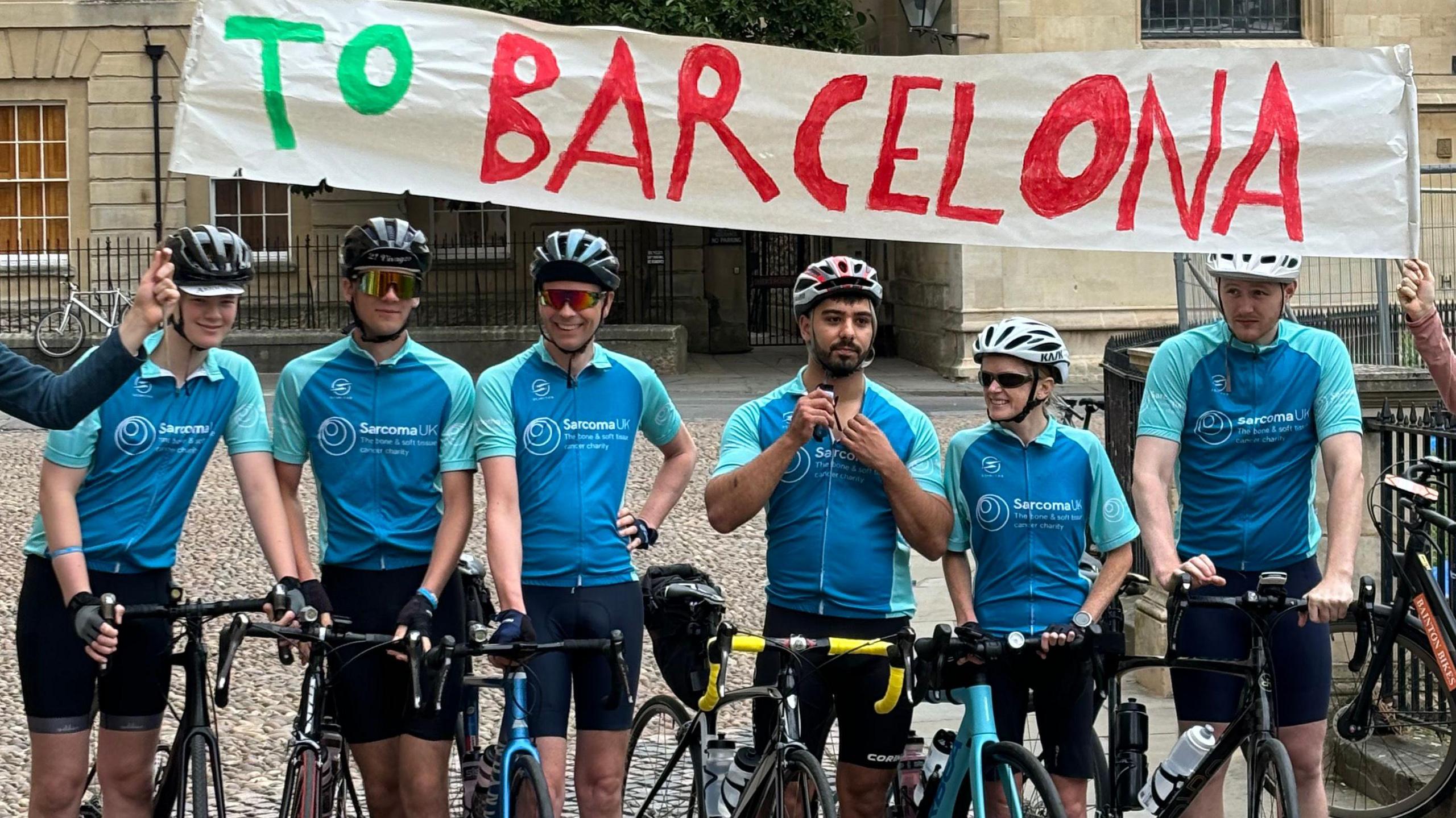 Six cyclists line up at the starting line at the Radcliffe Camera, including Dr Furniss and Jasper. A 'To Barcelona' banner is held above their heads.