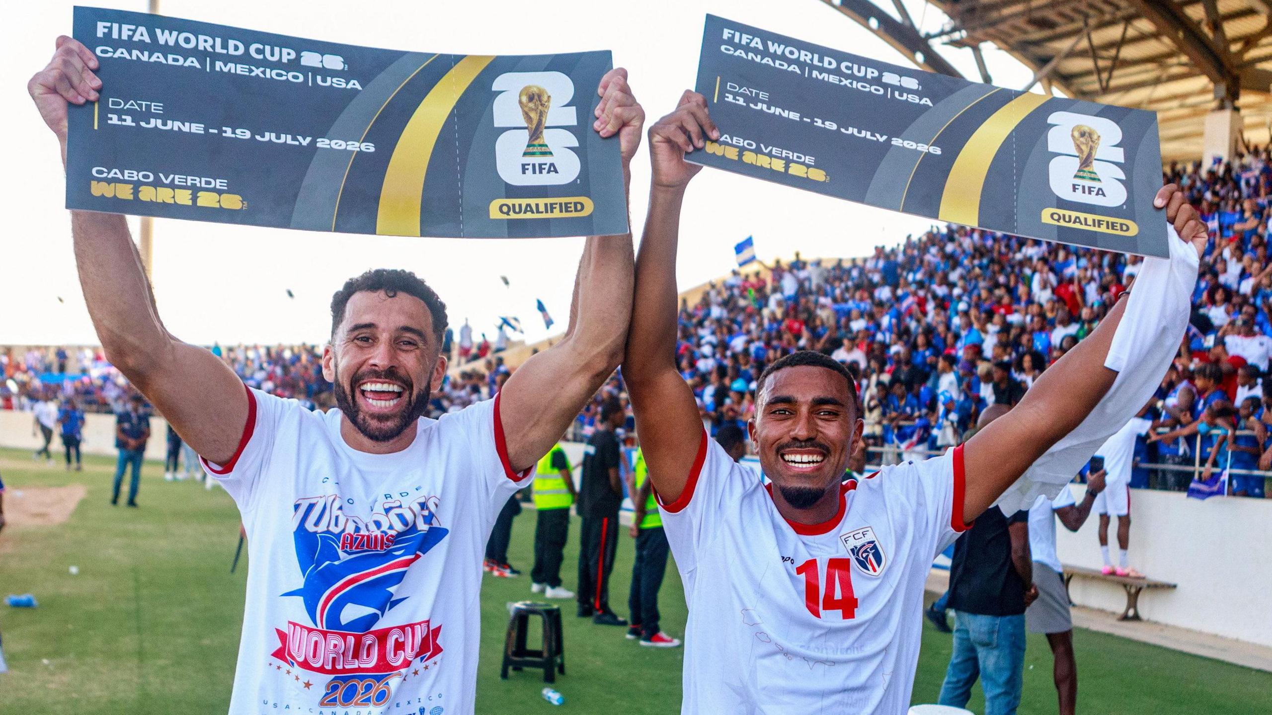 Roberto Lopes and Deroy Duarte, wearing white t-shirts, hold aloft banners detailing Cape Verde's qualification for the 2026 Fifa World Cup. In the distance behind them is a stand packed with Cape Verde fans