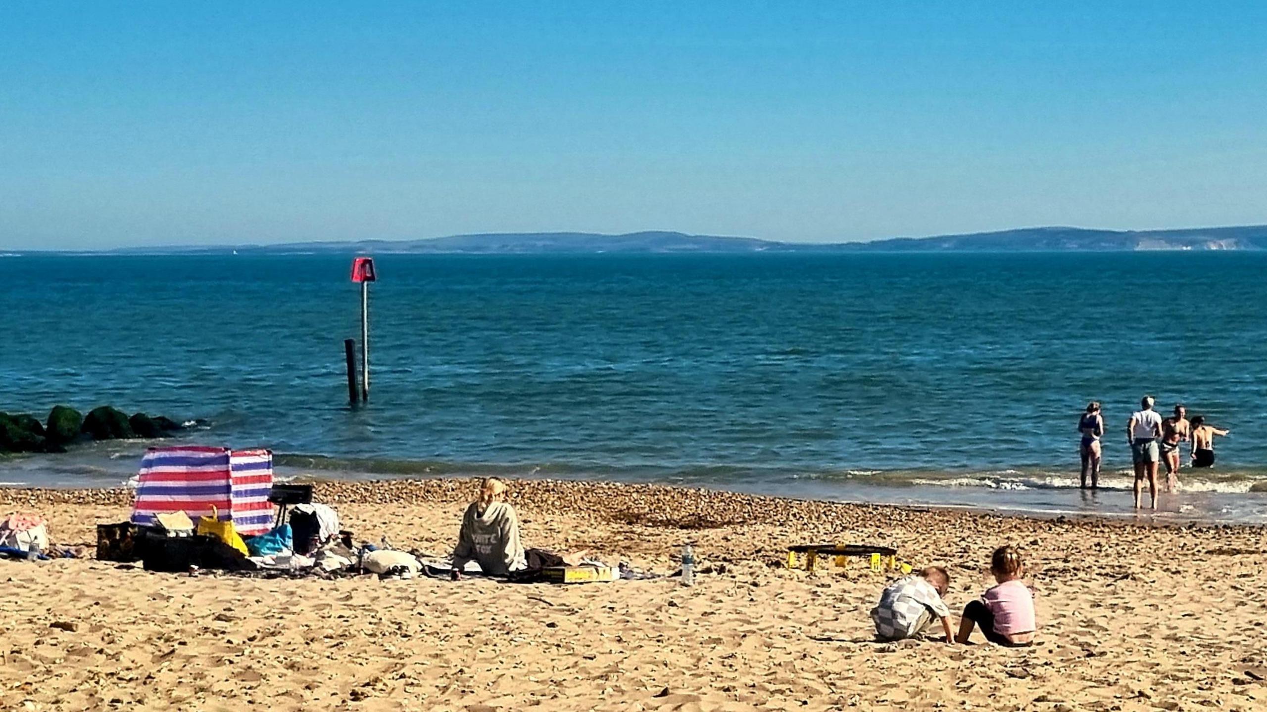 A beach scene with some people sat on the sand and a few others venturing out into the water. The blue sky is clear with hills in the background beyond the water.